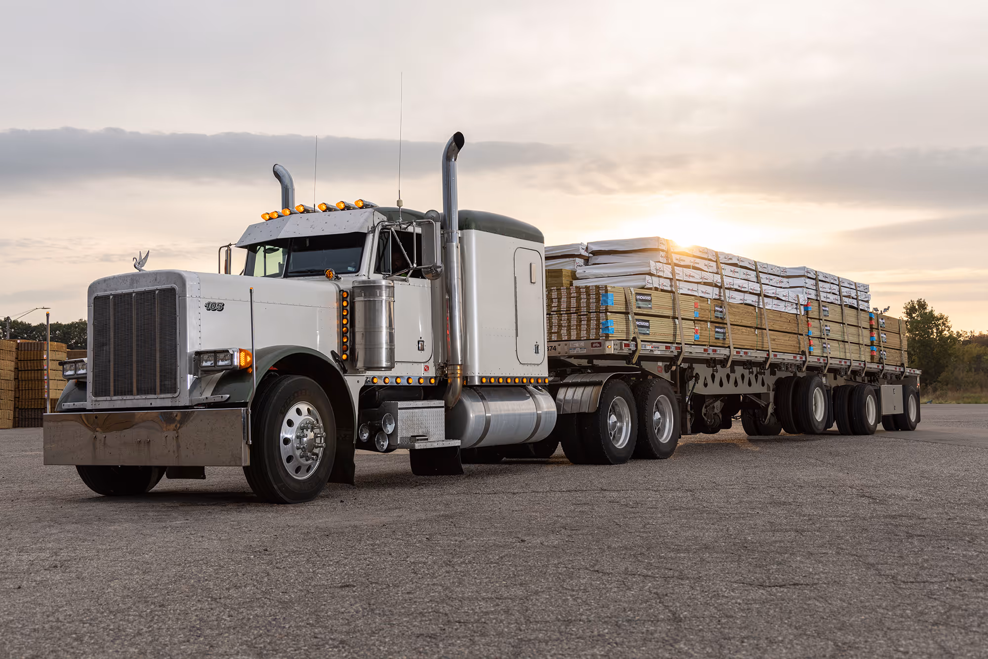 White semi-truck with a flatbed trailer loaded with stacked ProWood lumber and Deckorators decking at sunset.