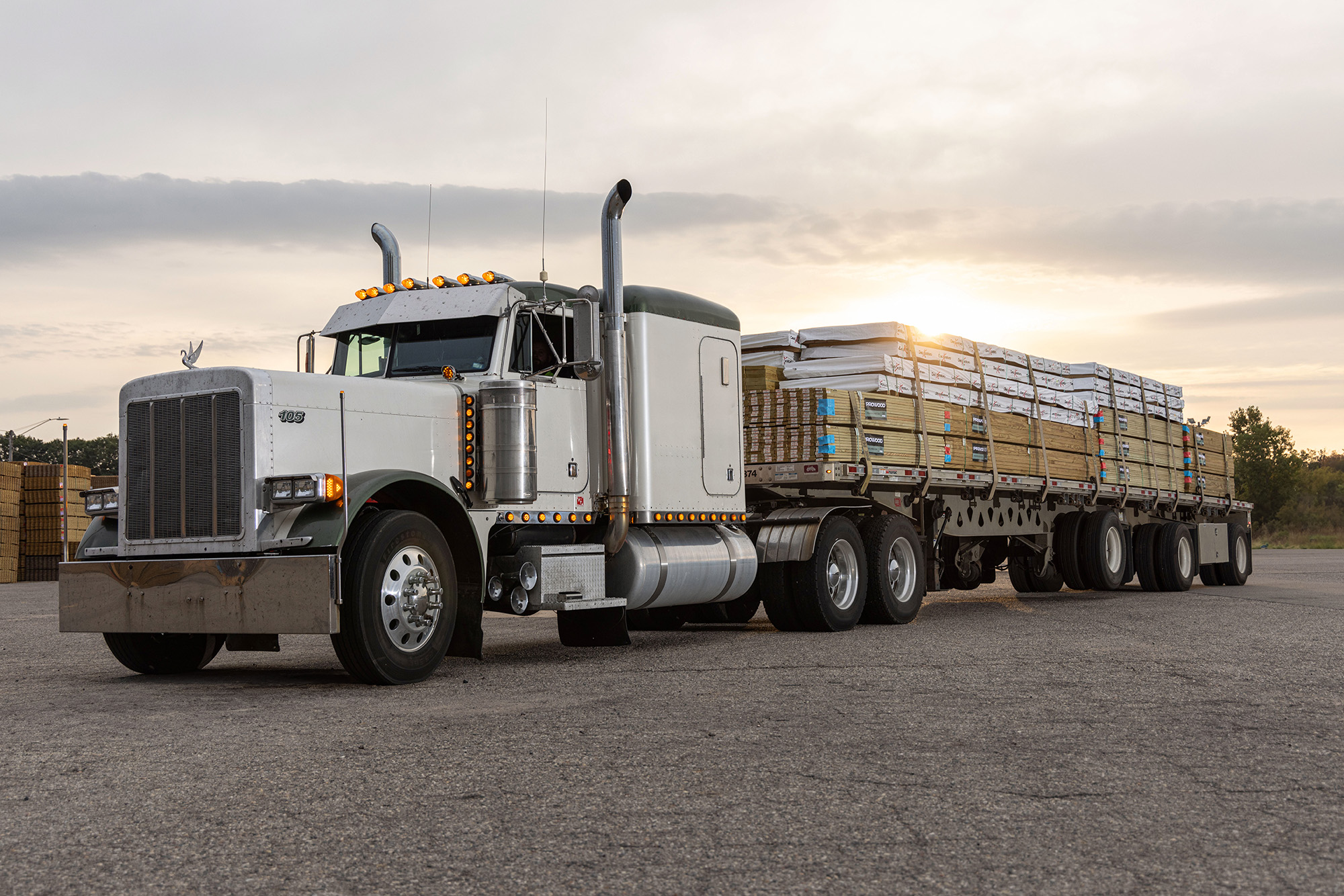 White semi-truck with a flatbed trailer loaded with stacked ProWood lumber and Deckorators decking at sunset.
