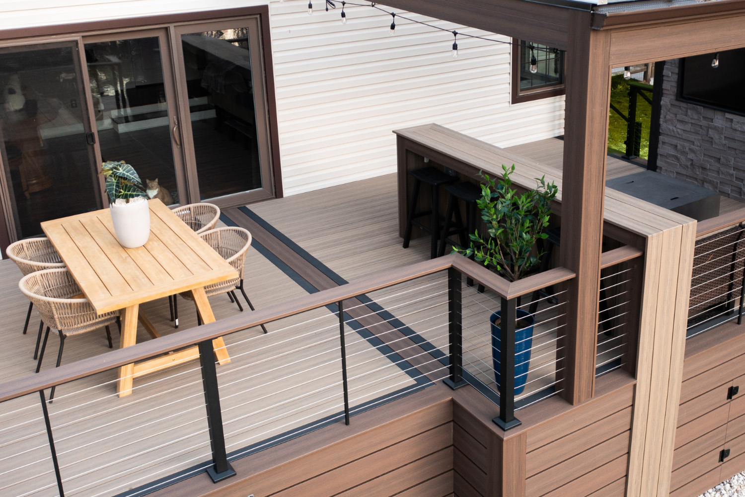 Outdoor deck with wood-look composite flooring, a wooden table with four wicker chairs, a potted plant on the table, a railing with metal cables, and a built-in bar area with stools and string lights.