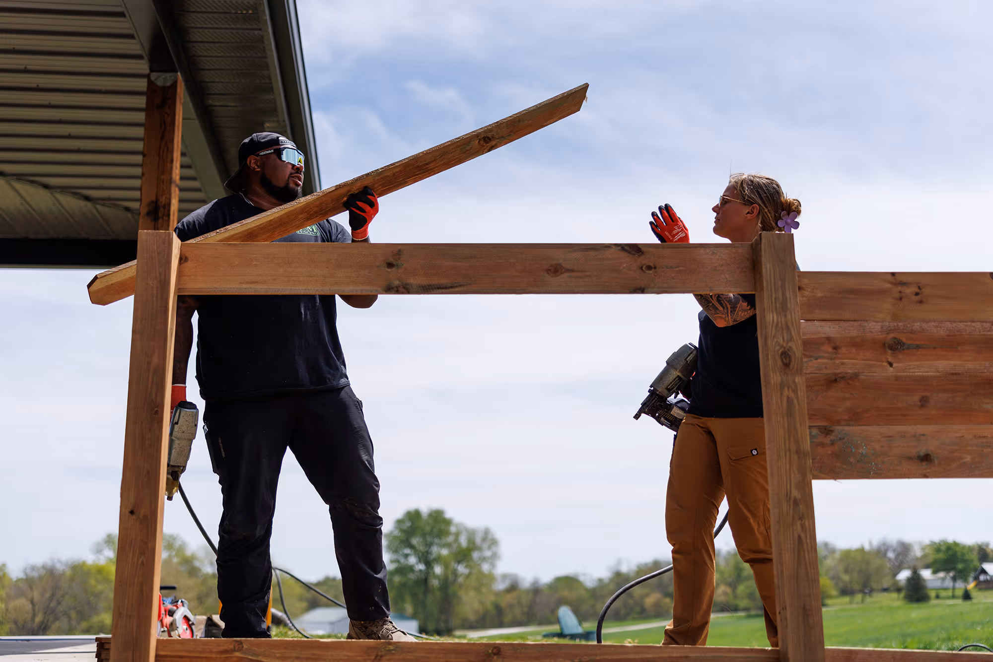 Two construction workers building a wooden fence outdoors, one holding a wooden plank and the other a nail gun.