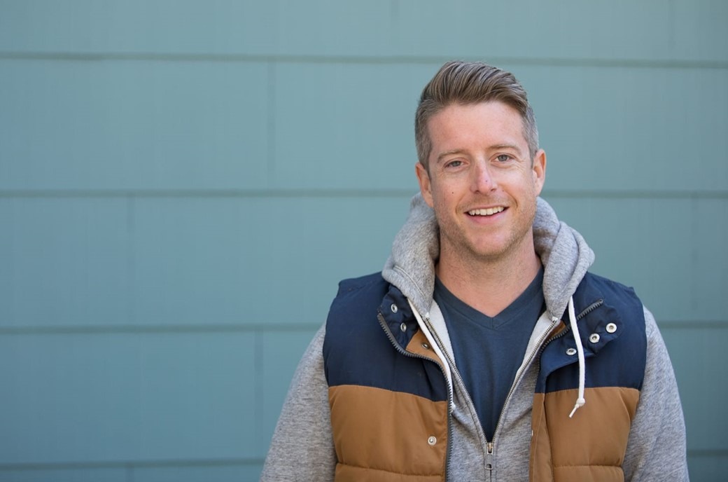 Smiling man wearing a blue and brown vest over a gray hoodie standing in front of a teal paneled wall.
