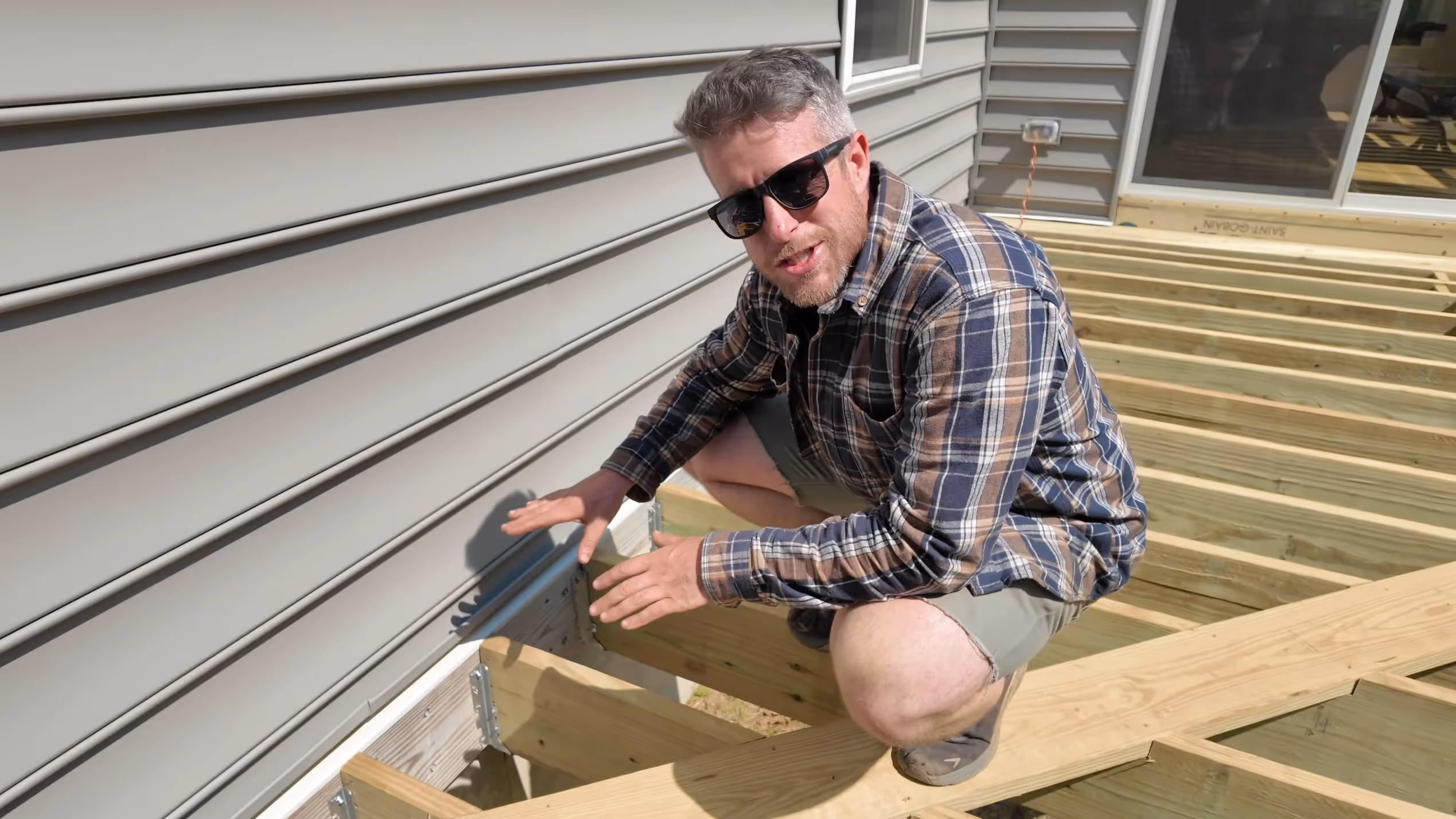 Man in sunglasses and plaid shirt demonstrating wooden deck framing against a house wall.
