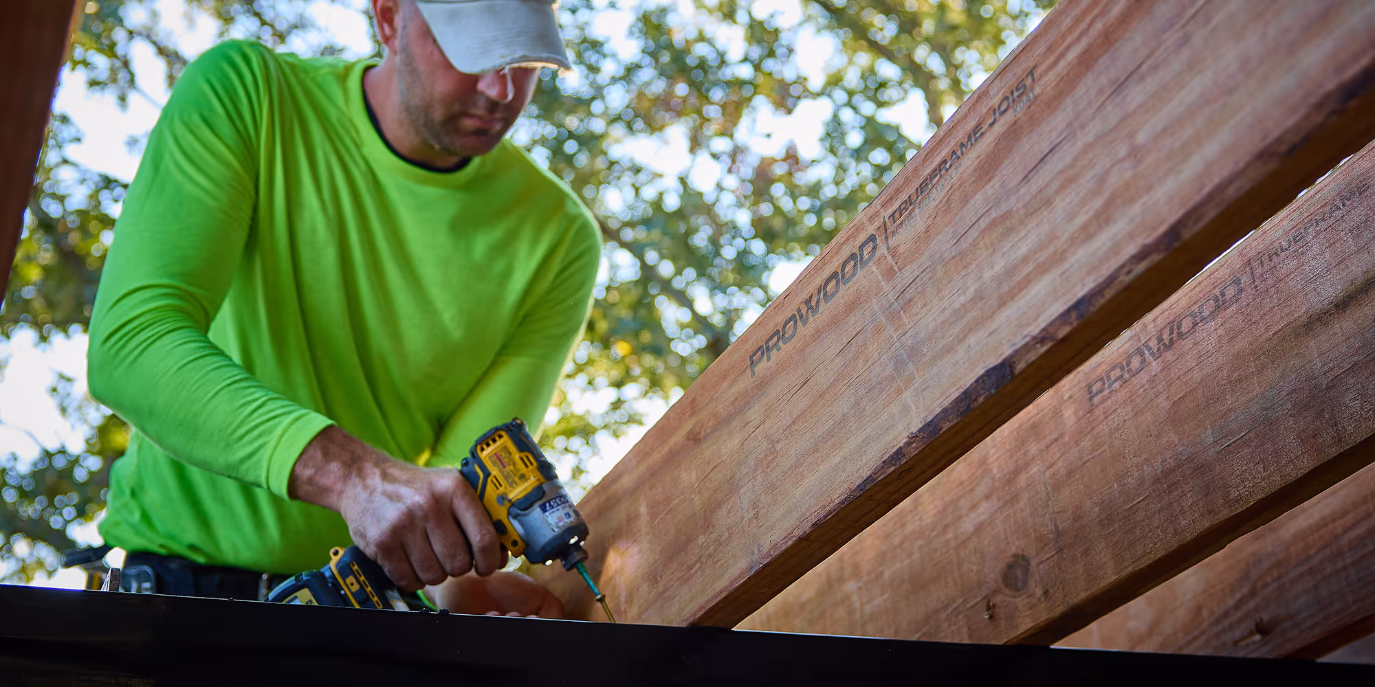 Construction worker in a bright green shirt using a power drill to fasten wooden beams labeled ProWood.