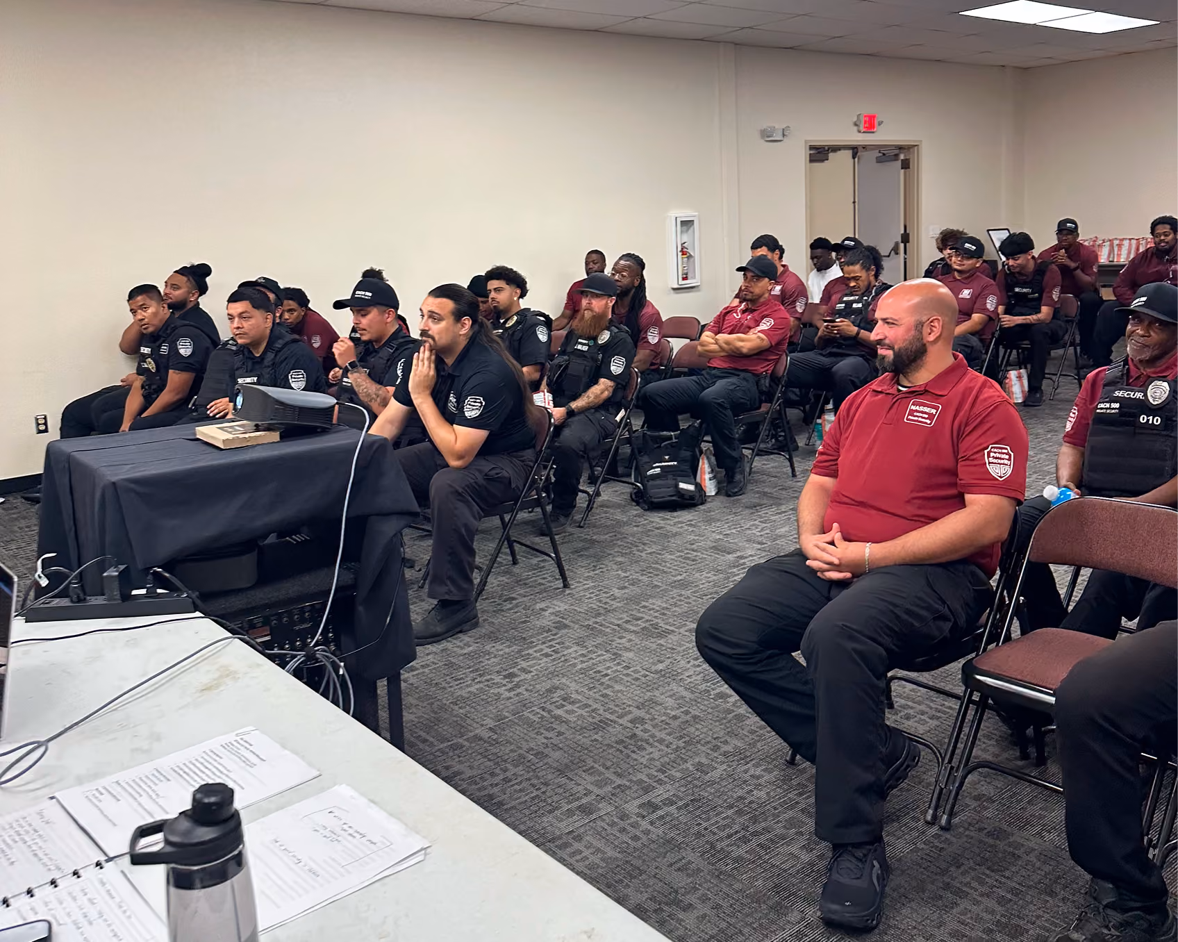 A group of security personnel in black and red uniforms seated attending a training session in a classroom.