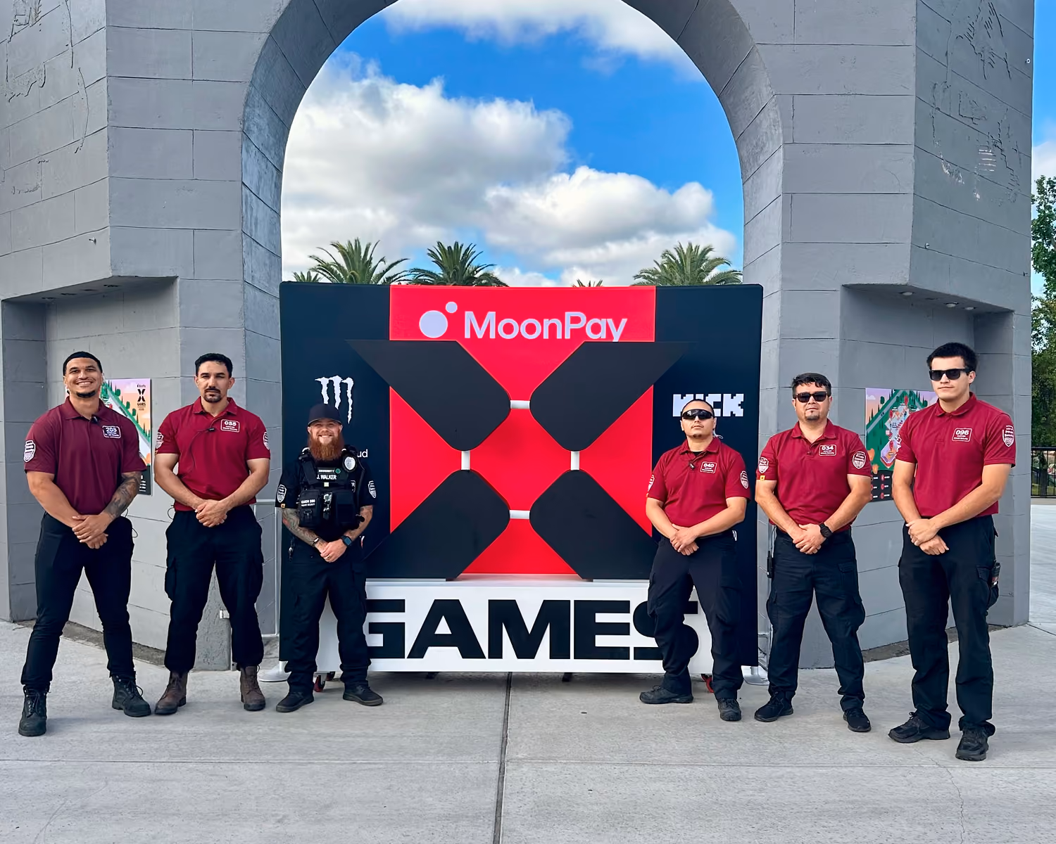 Six security guards standing in front of a large X Games sign under a gray arch outdoors.