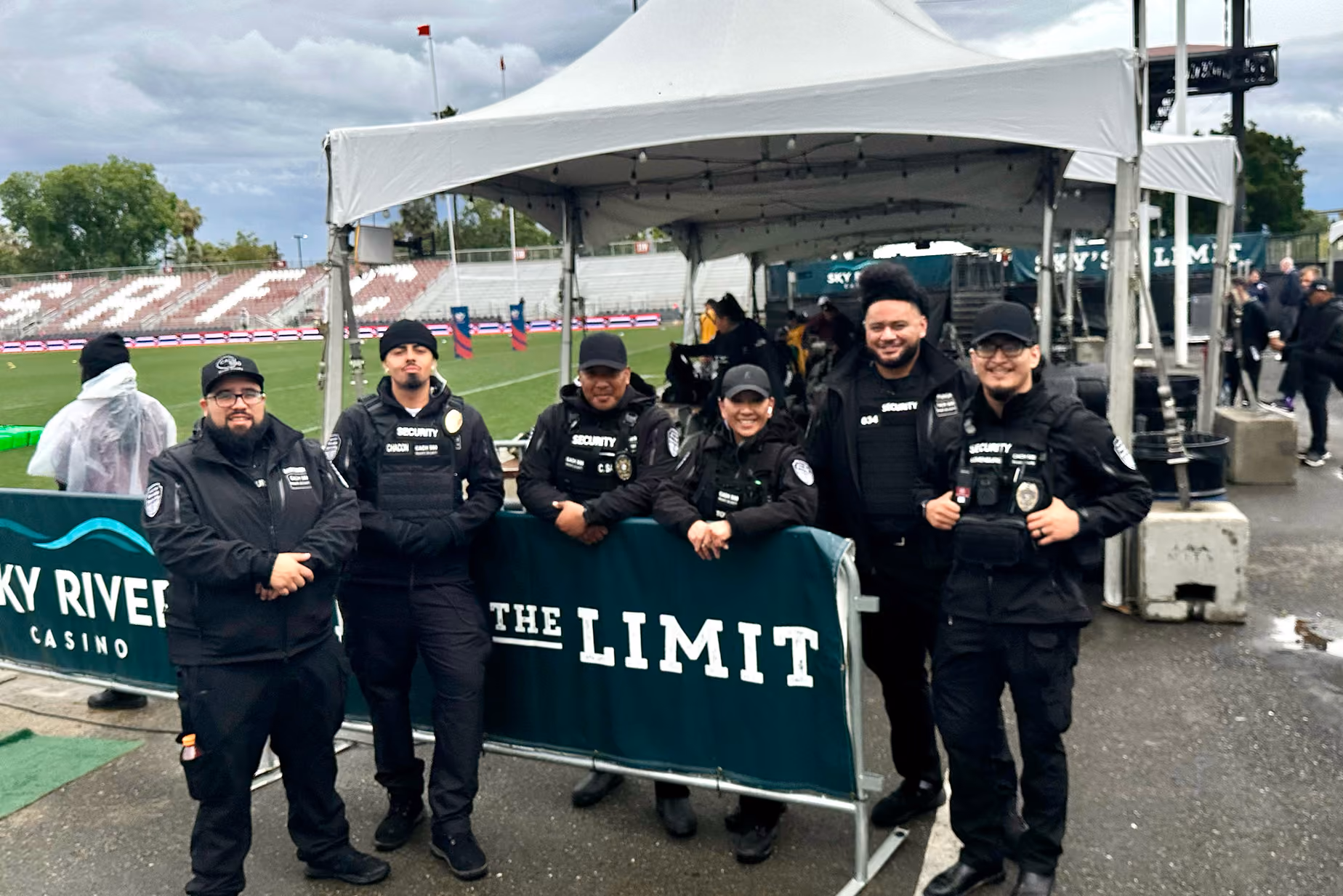Six security guards dressed in black uniforms and vests standing and leaning relaxedly by a green barrier with white text reading 'SKY RIVER CASINO THE LIMIT' under a white outdoor tent at a sports event.