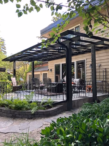Outdoor patio with black metal pergola, surrounded by plants and garden, attached to a beige house.