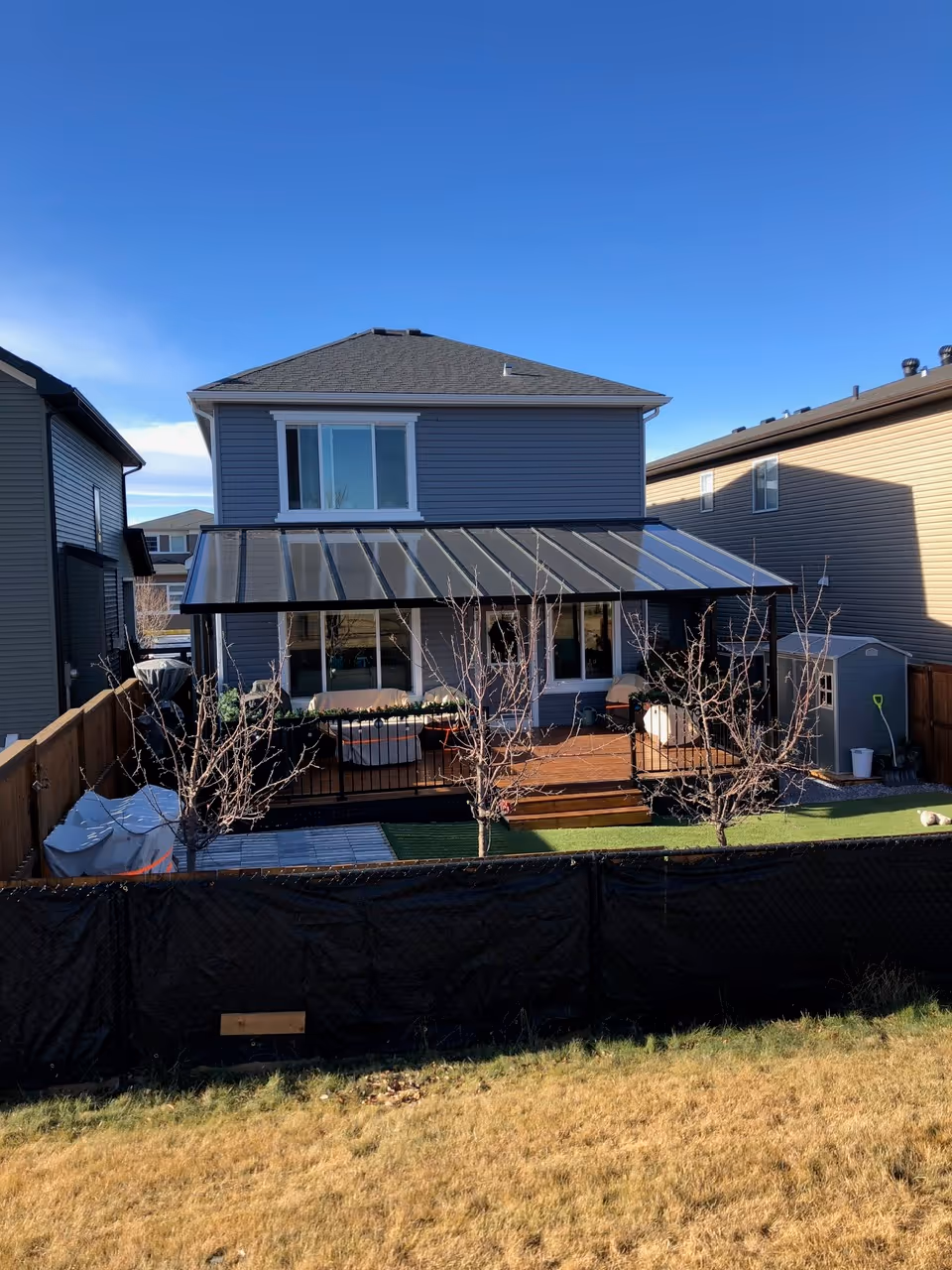 Backyard view of a two-story gray house with a covered wooden deck, leafless trees, and a shed on the right side under a clear blue sky.