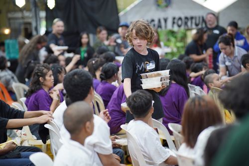 A young boy handing out food to a group of young people and the elderly.