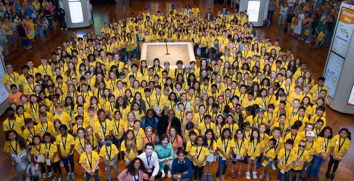 A high level picture of a huge room filled with hundreds of kids all wearing a yellow invention convention shirt