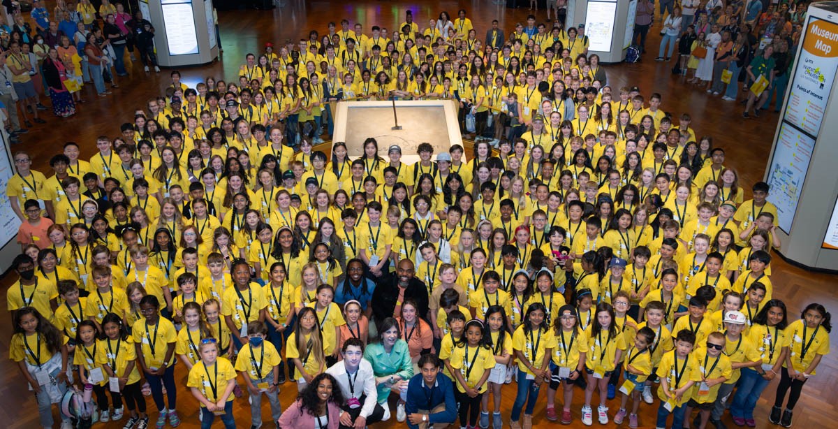 A high level picture of a huge room filled with hundreds of kids all wearing a yellow invention convention shirt