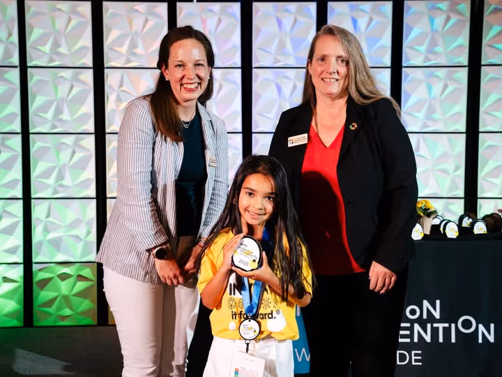 Two women smiling while standing around a smiling little girl who is holding up her Invention Convention medals