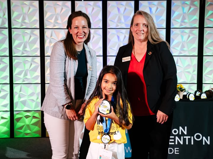 Two women smiling while standing around a smiling little girl who is holding up her Invention Convention medals