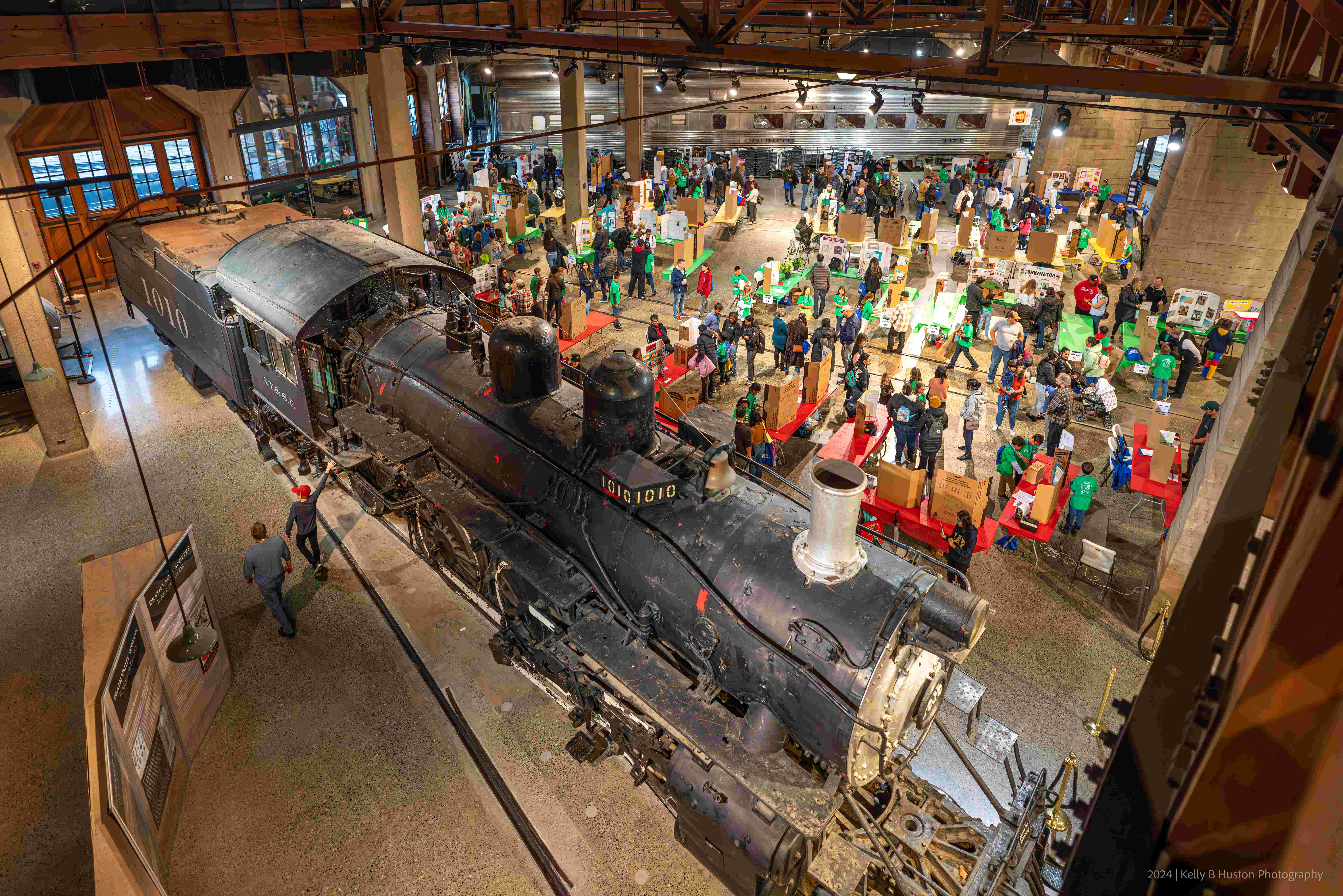 Over the top picture of the Sacramento Raiload Museum, featuring a big black train with many museum visitors seen behind it.