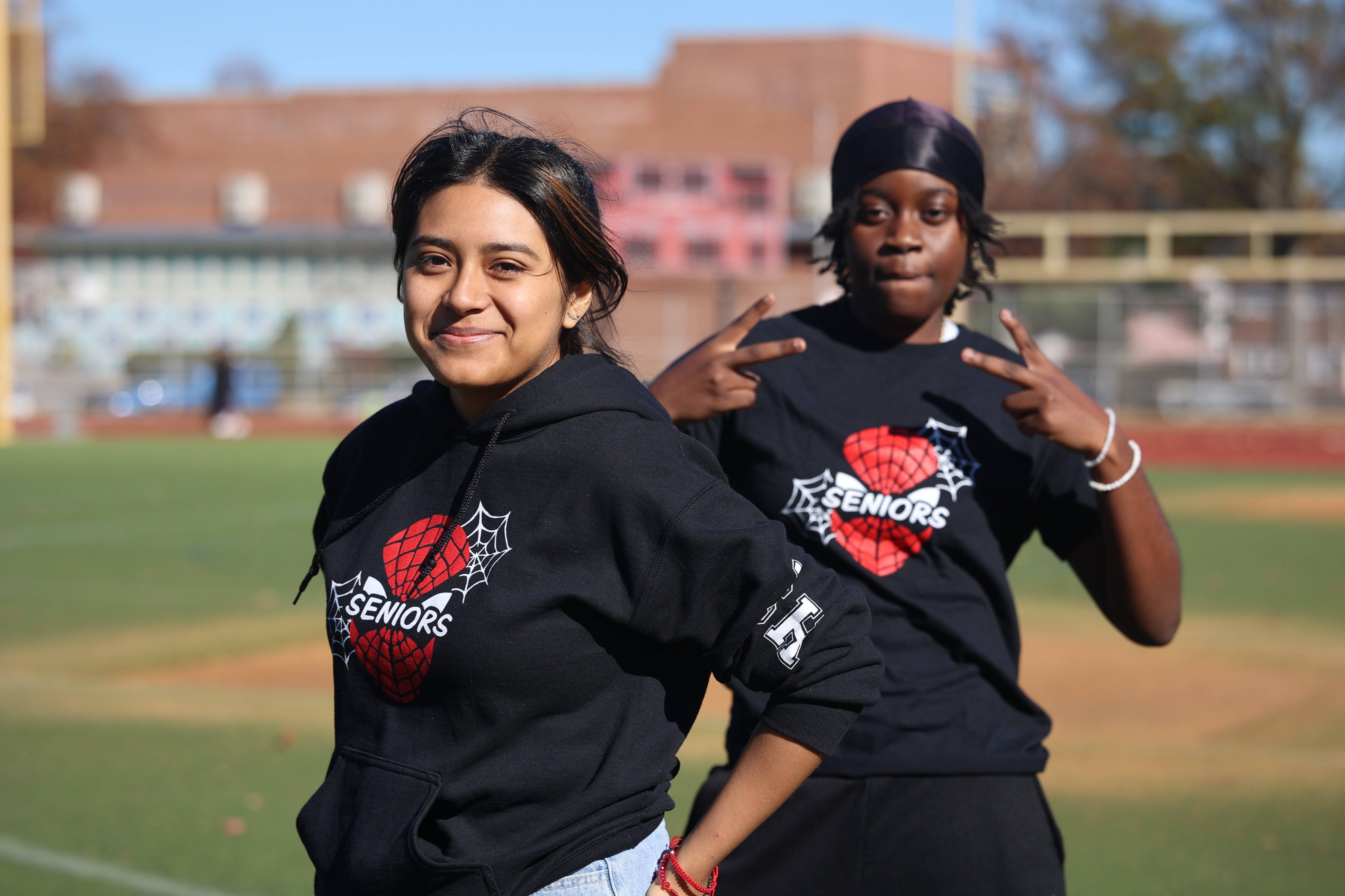 Two high school seniors wearing matching black Seniors hoodies on athletic field