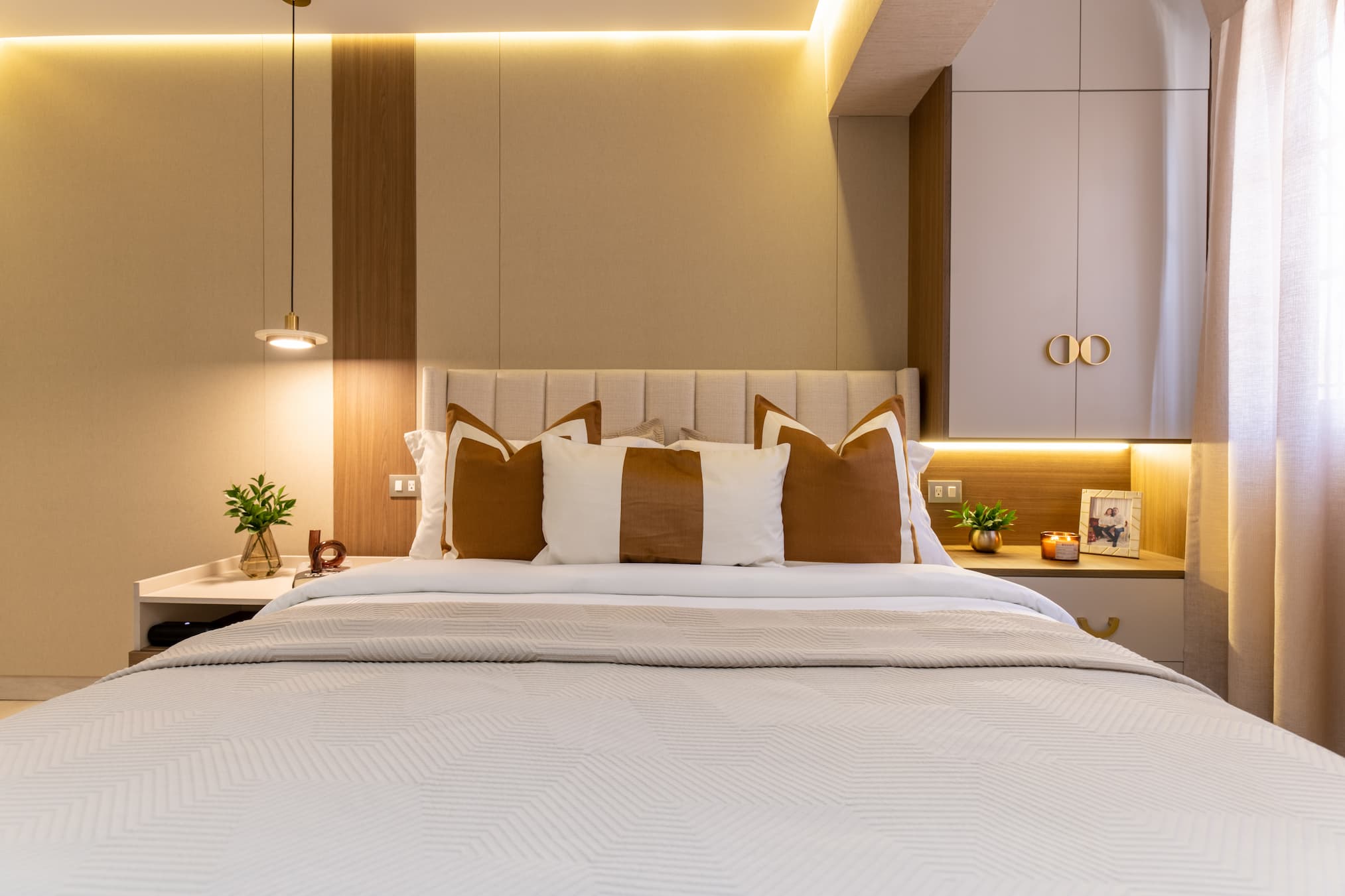 Chambre moderne avec lit double décoré de coussins blancs et marron, lampes suspendues, et tables de chevet avec plantes, bougie et photo encadrée. Photographie Immobilier Hôtel Chambre lit double deluxe - Arkenyte