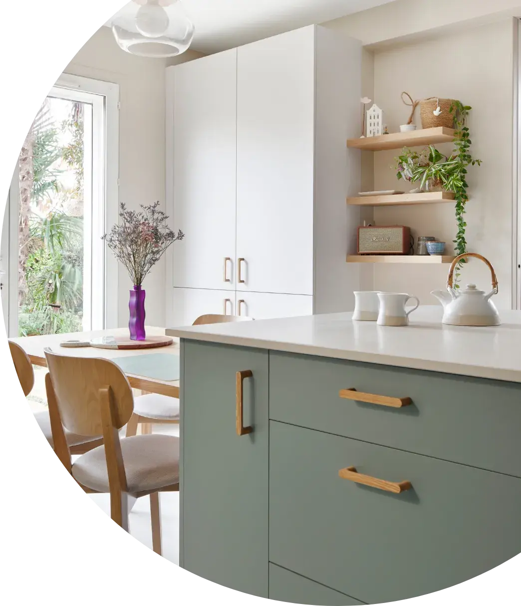 Dès les premiers échanges, leurs envies étaient très claires : une cuisine bicolore lin ivoire et vert eucalyptus, réchauffée par des touches de bois, et sublimée par un plan de travail en céramique. Si l’esthétique était déjà bien définie, le projet a également intégré des éléments sur-mesure, notamment une colonne de réfrigérateur pensée pour offrir un véritable espace de rangement au-dessus du frigo, sans contrainte d’ouverture. Semi-ouverte sur le salon, cette cuisine est une pièce à part entière. Baignée de lumière naturelle grâce à une ouverture vers l’extérieur, elle s’intègre parfaitement à l’espace de vie.