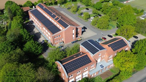 Luftaufnahme von Gebäuden mit Solarpaneelen auf den Dächern und umgeben von Bäumen und Parkplätzen.