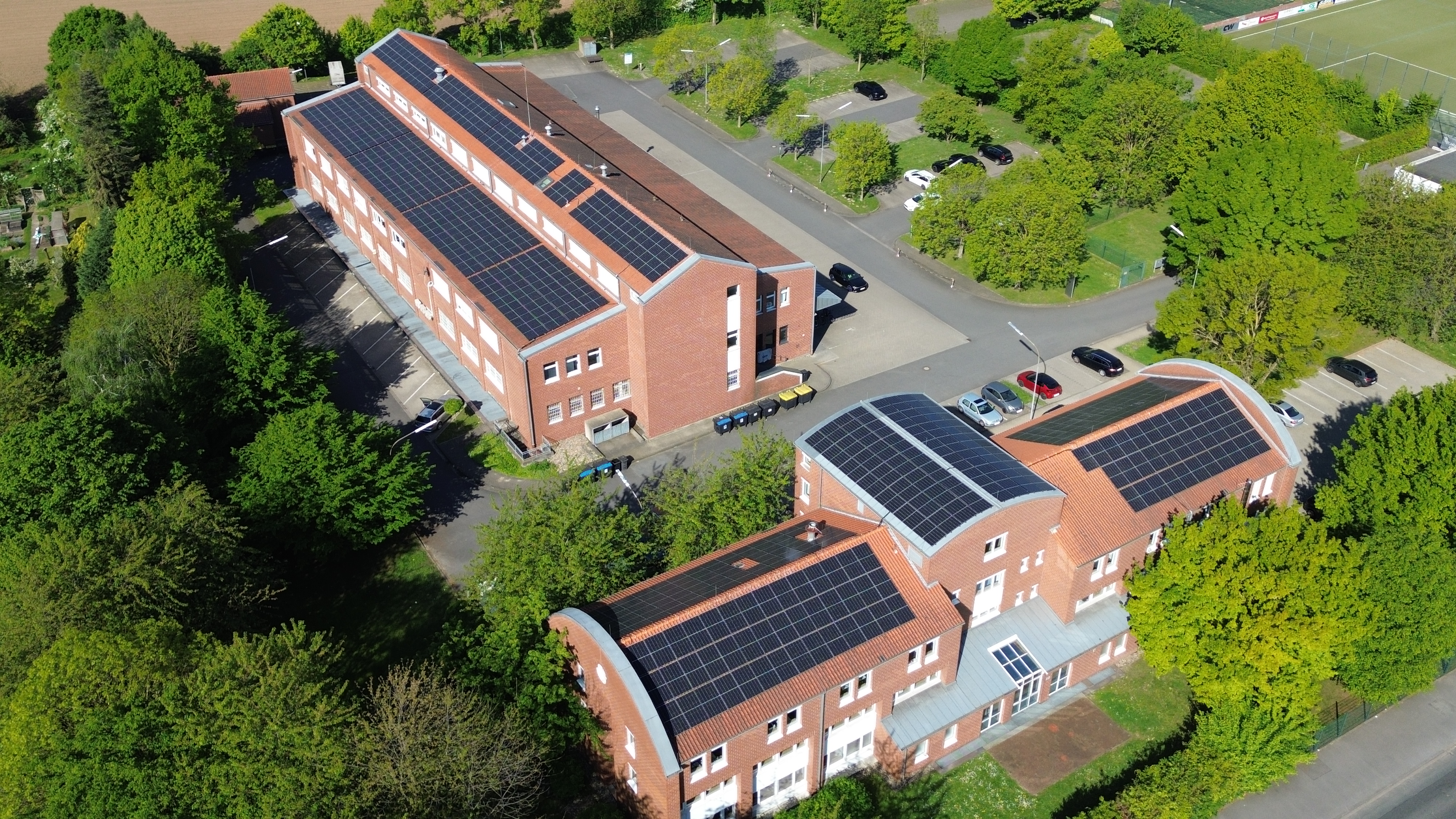 Luftaufnahme von Gebäuden mit Solarpaneelen auf den Dächern und umgeben von Bäumen und Parkplätzen.