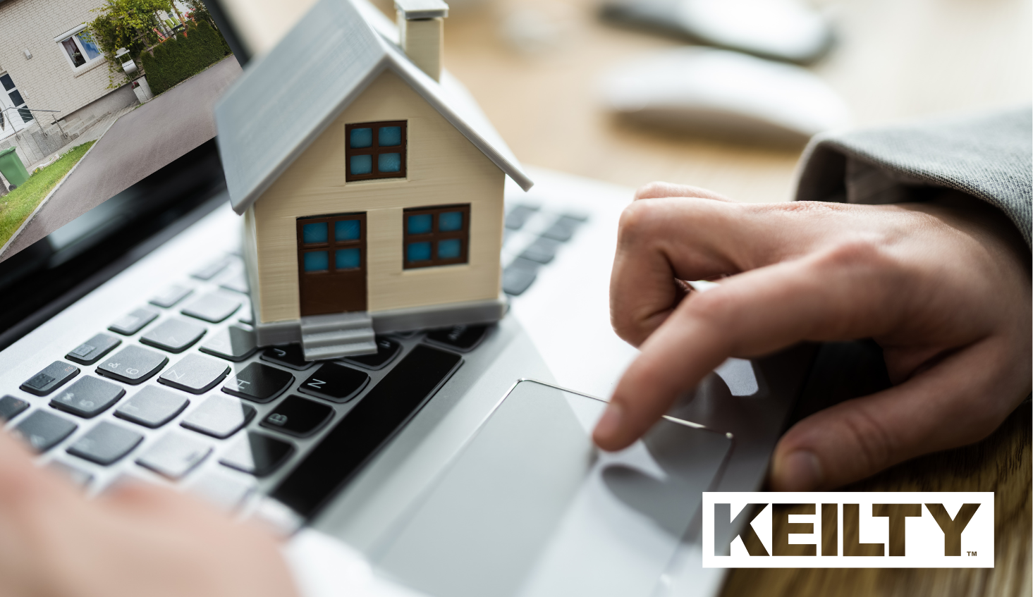 A model wooden house sitting on a laptop keyboard while a hand uses the trackpad, illustrating the KEILTY owner portal.