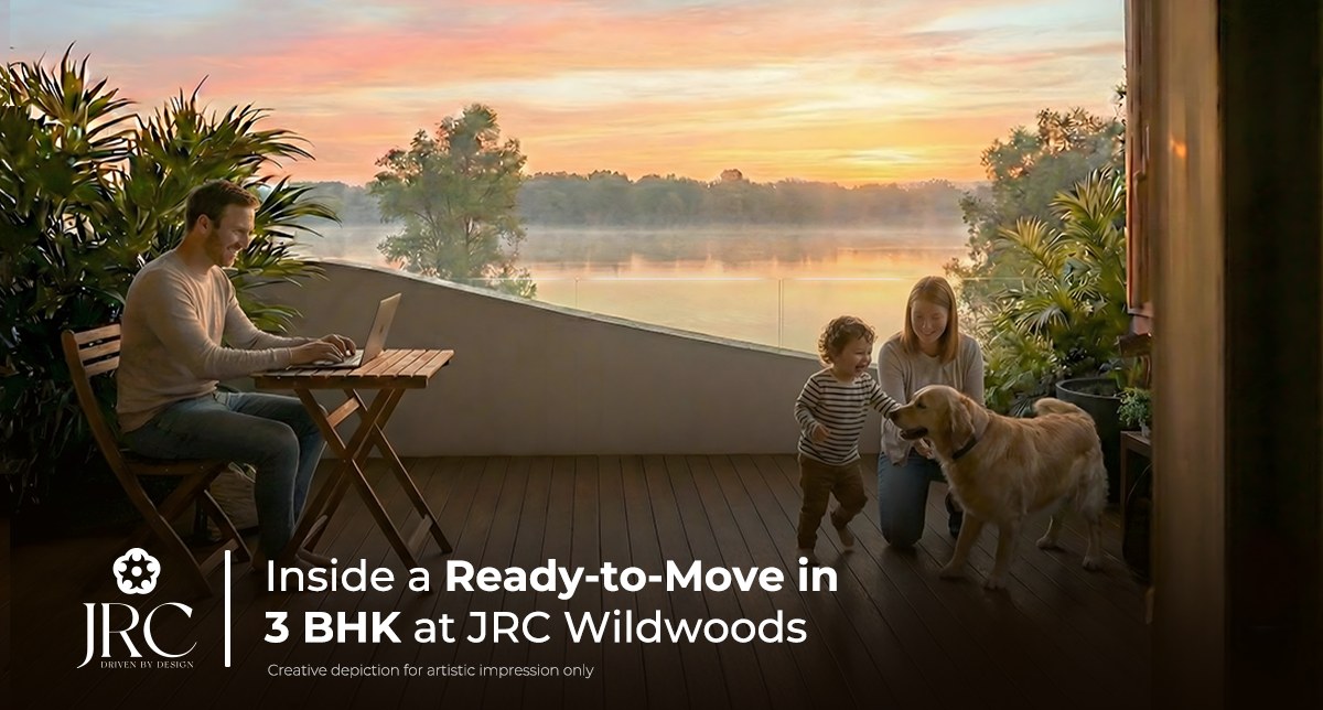 Family on the balcony of a ready-to-move 3 BHK lakefront apartment at JRC Wildwoods off Sarjapur Road, Bangalore — one parent working on a laptop while a child and dog play, with mature tree canopy and the lake beyond the rail at sunrise