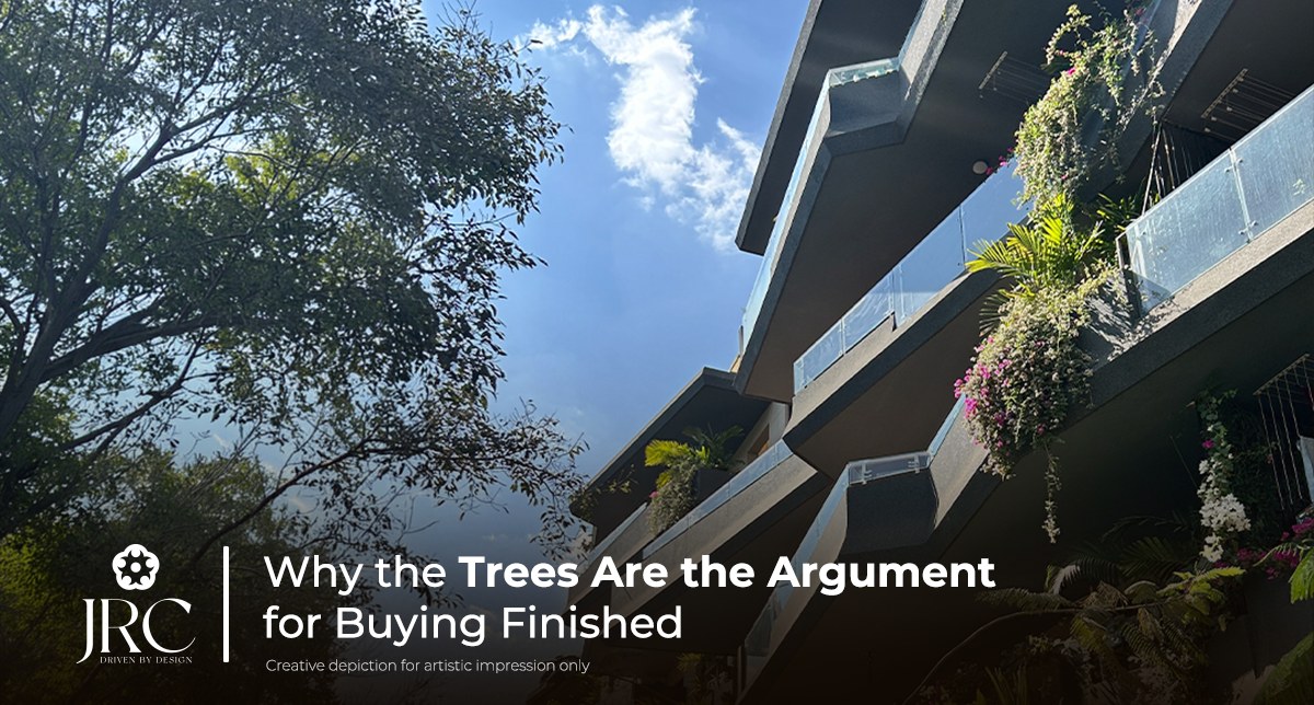 Upward view of tree-hugging balconies at JRC Wildwoods against open sky, with a mature tree in the foreground and bougainvillea spilling over the rails — a ready-to-move low-density apartment community off Sarjapur Road, Bangalore