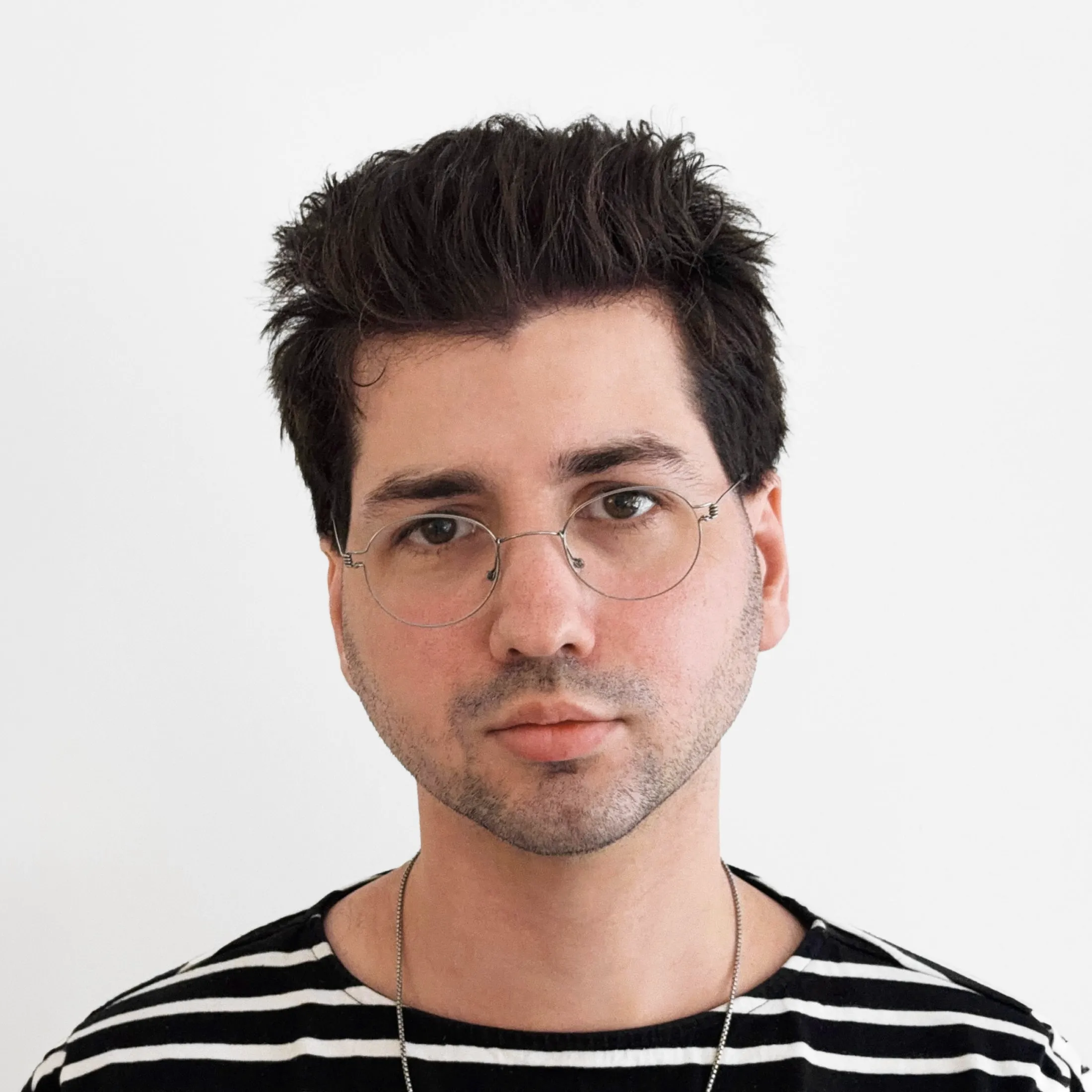 Portrait of Dylan Seeger with dark, tousled hair, wearing round wireframe glasses, a black and white striped shirt, and a silver chain necklace, against a plain white background.