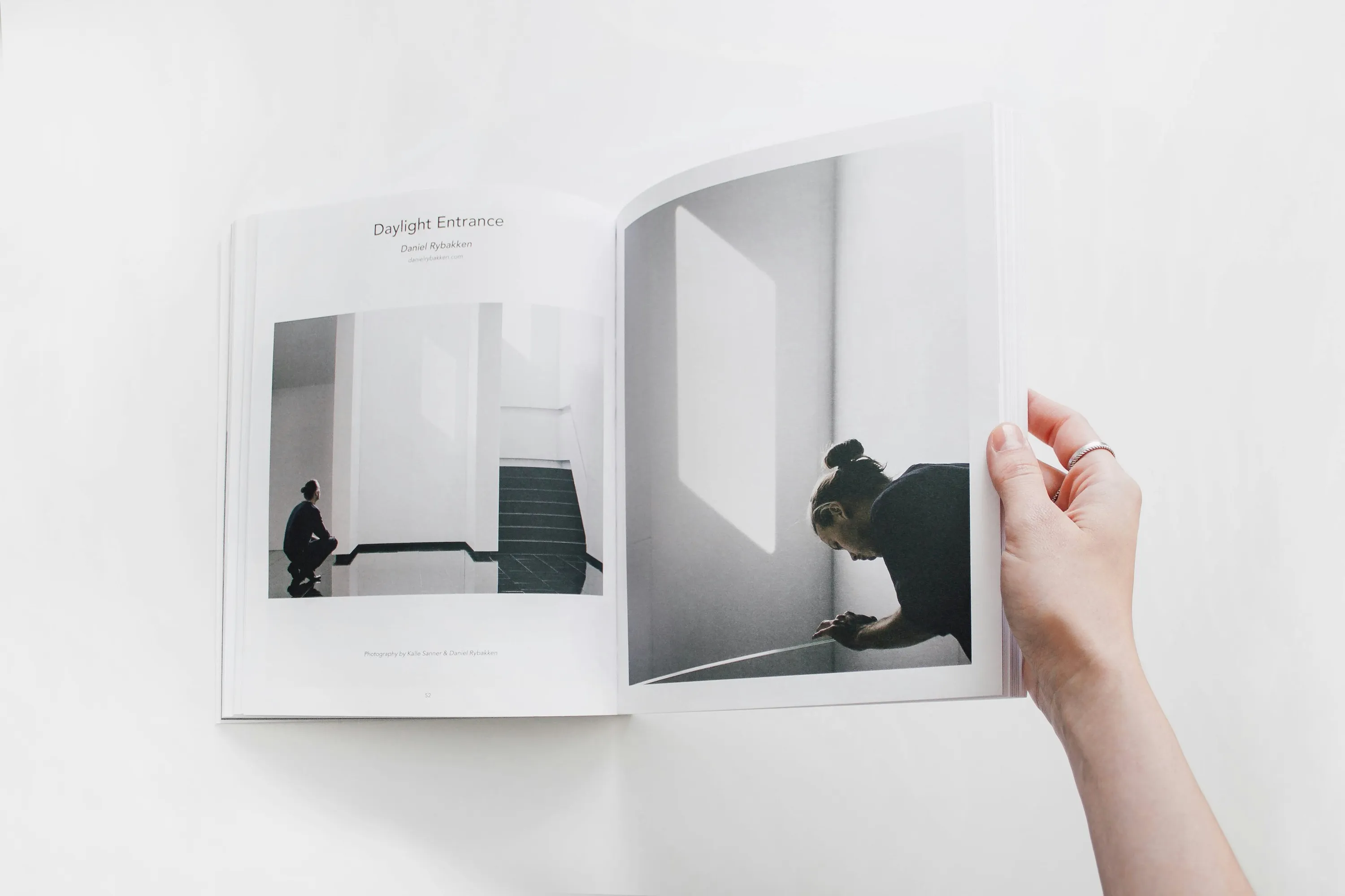 Hand holding an open book showing two photographs of a person in dark clothing interacting with light and shadows in a minimal indoor space with white walls and stairs.