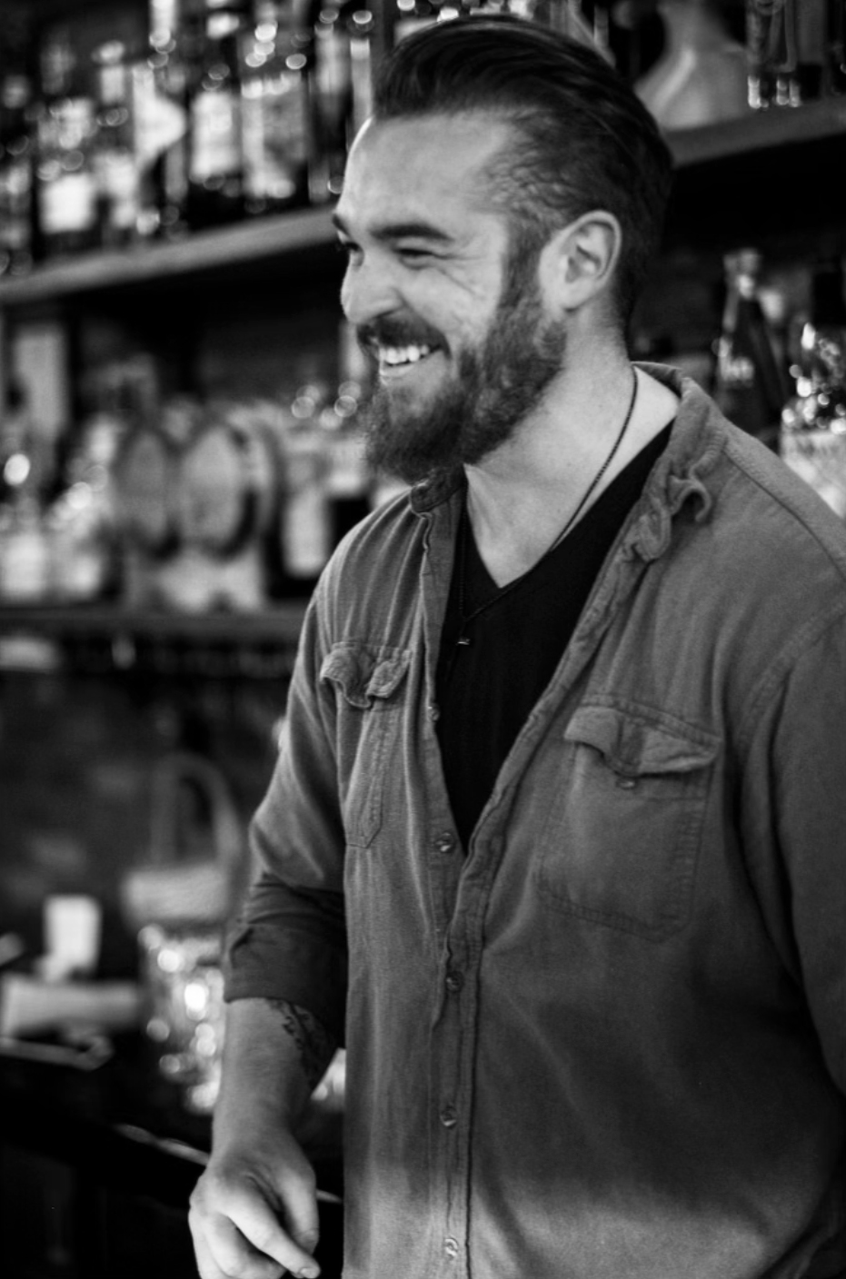 Bartender Austin Parsons smiling ear-to-ear with shelves full of a large array of liquor