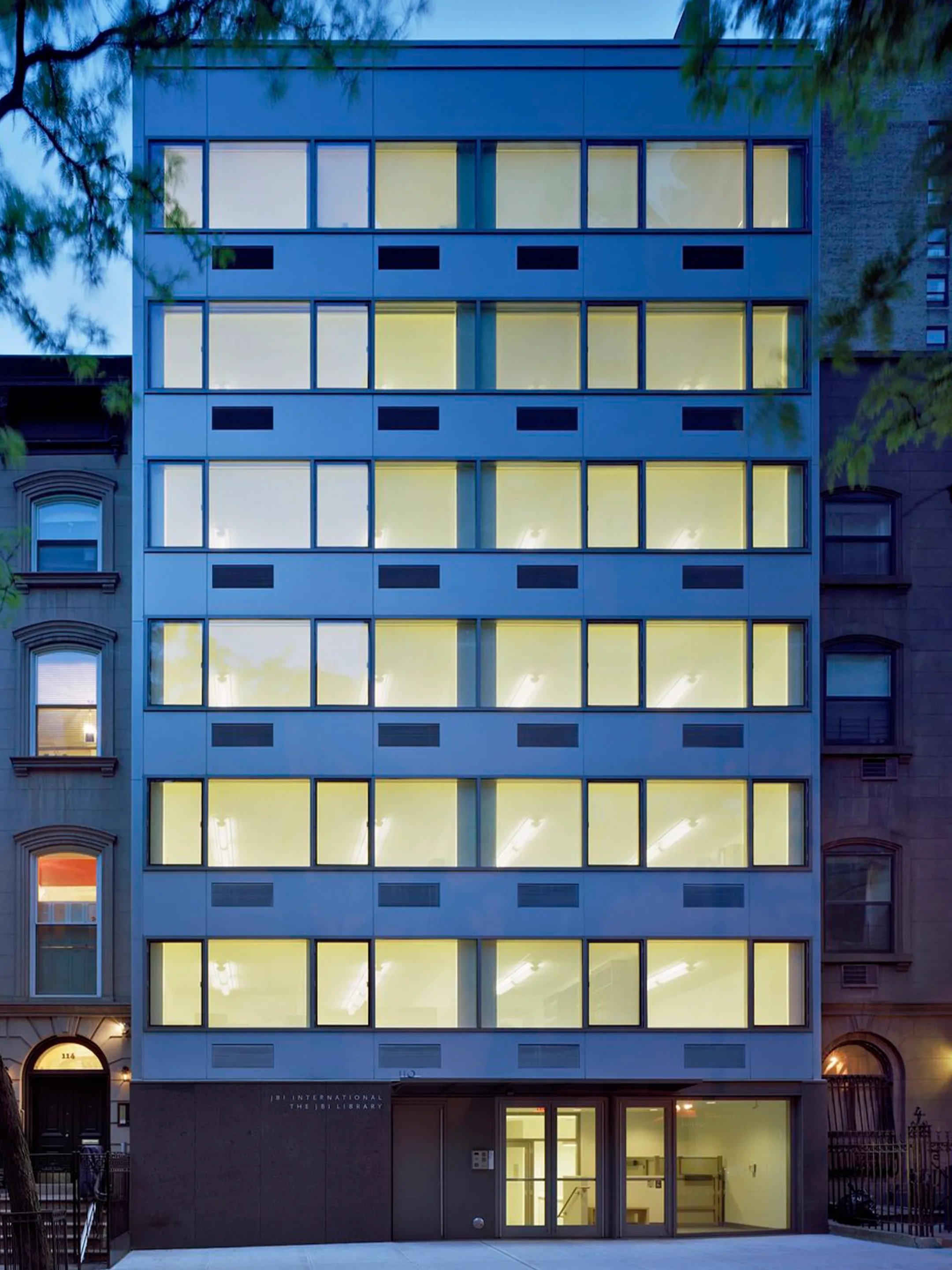 Modern building with rows of illuminated windows between older brick buildings at dusk.