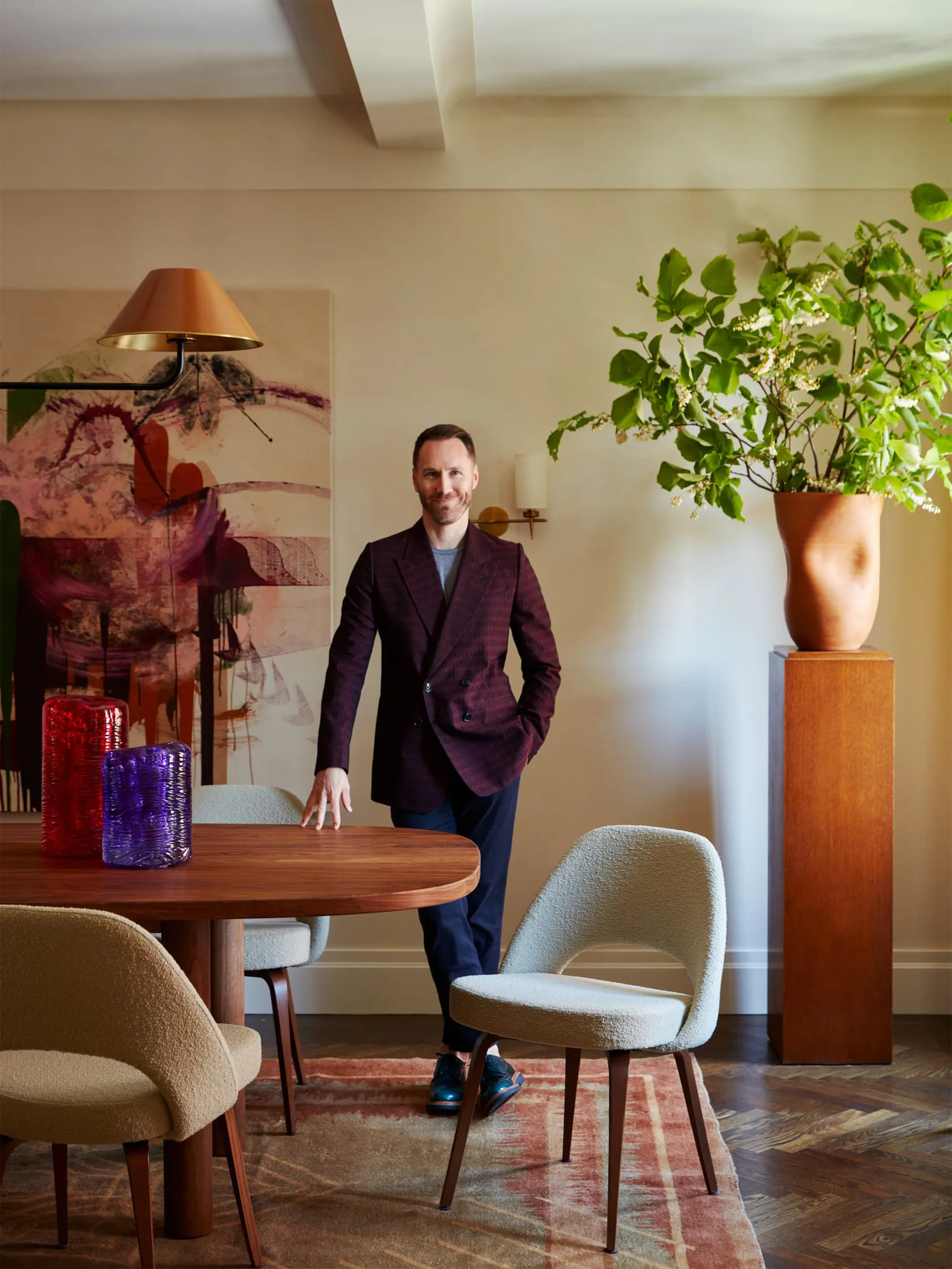 Josh Greene standing beside a round dining table in a room with abstract art and leafy branches.