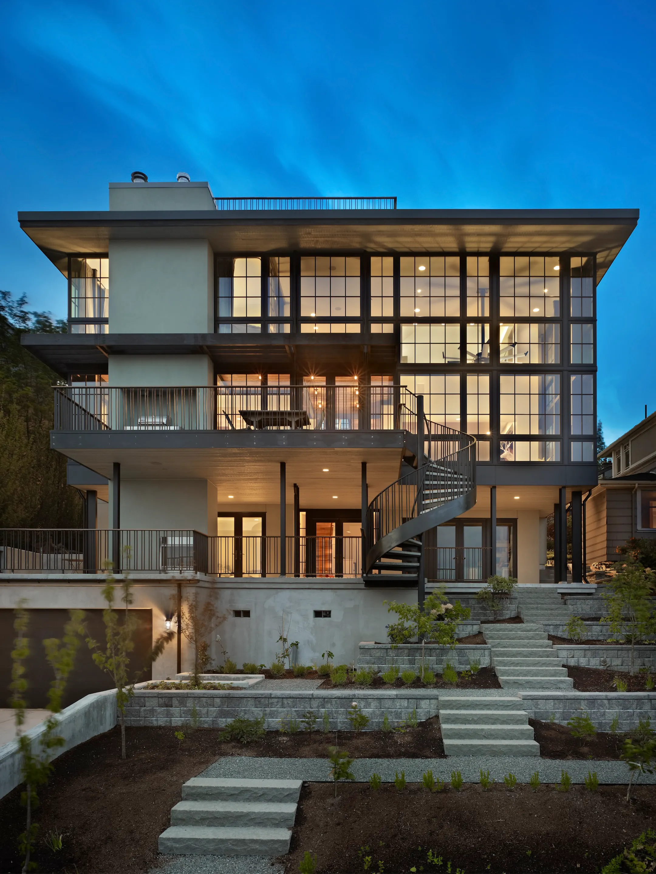 Modern house at dusk with tall illuminated windows, an exterior spiral stair, and terraced garden.