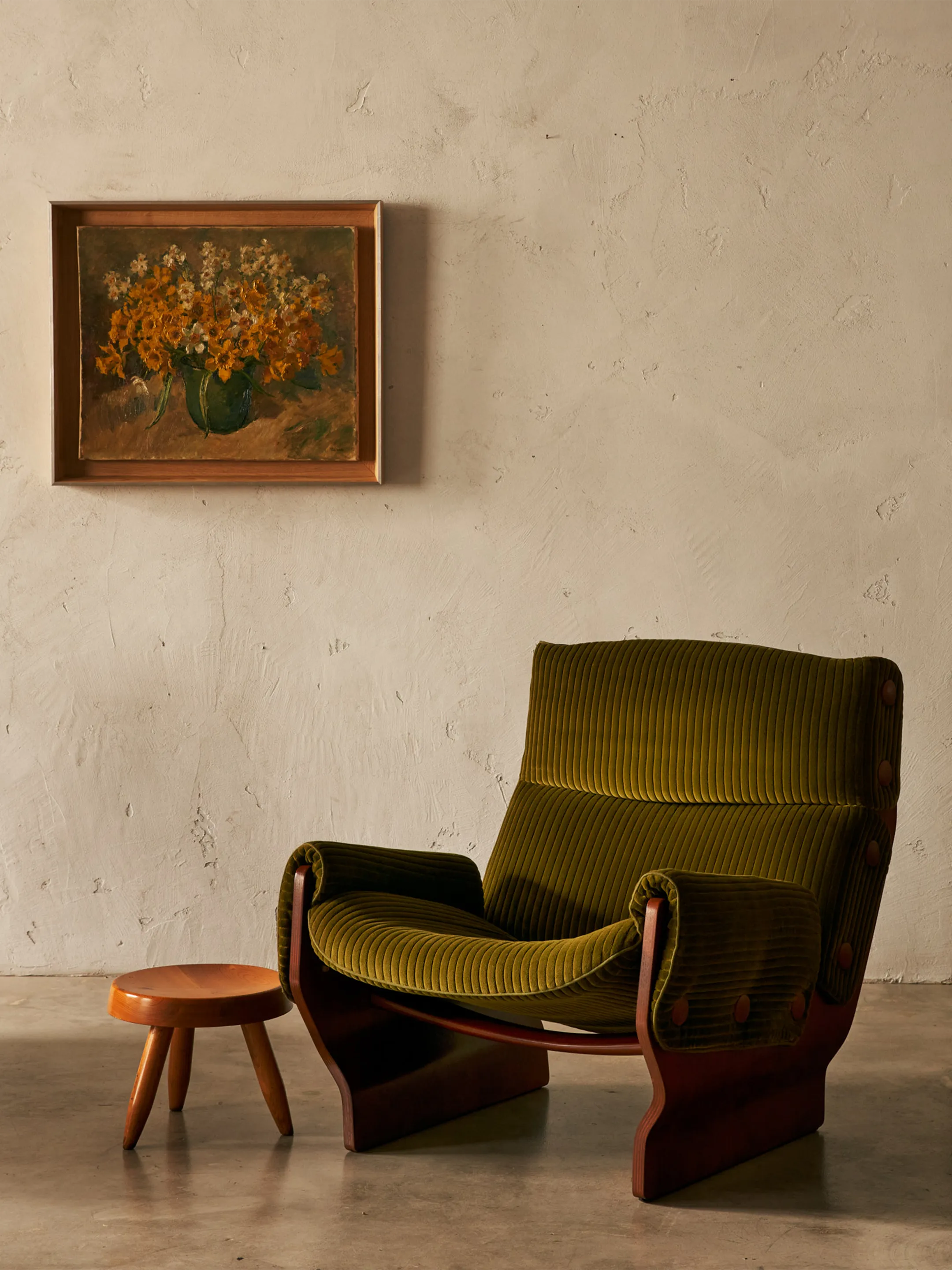 Olive-green upholstered armchair and small wooden stool beneath a floral painting.