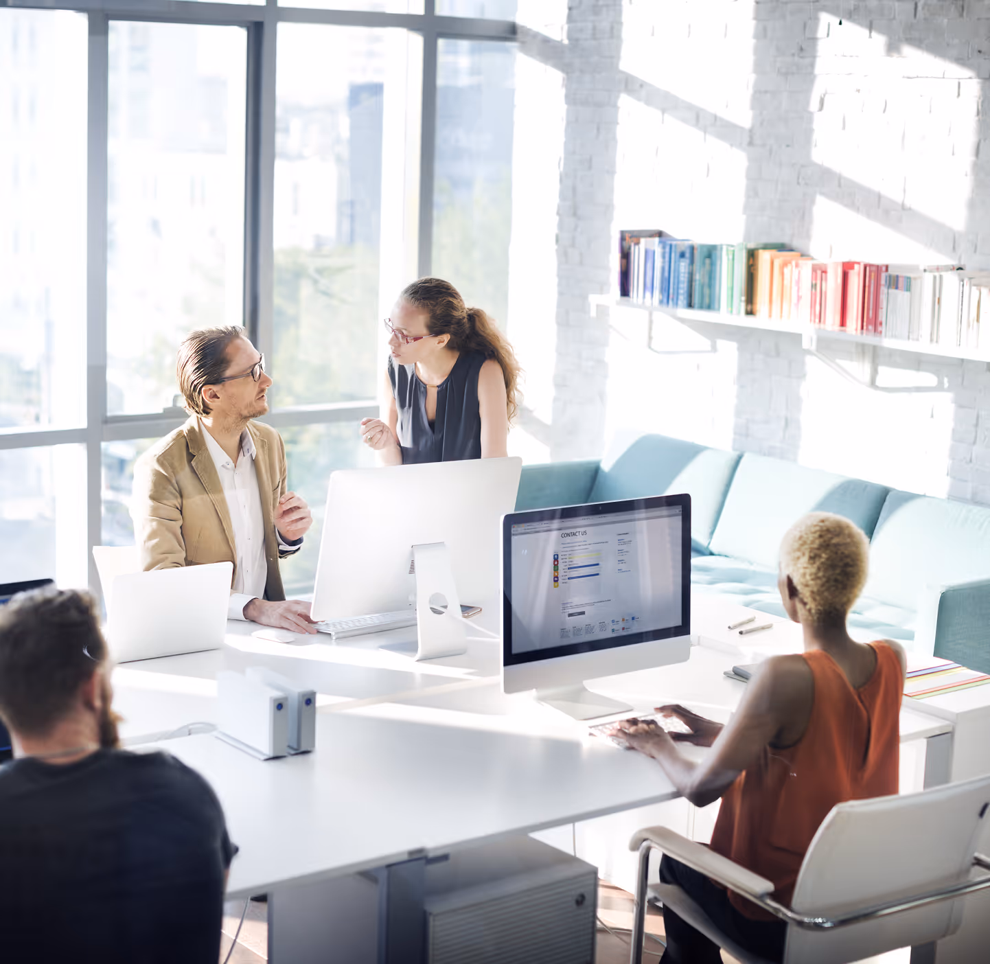 A group of people working together in a modern office. Two people are talking, while one sits across a desk and works on a desktop monitor.