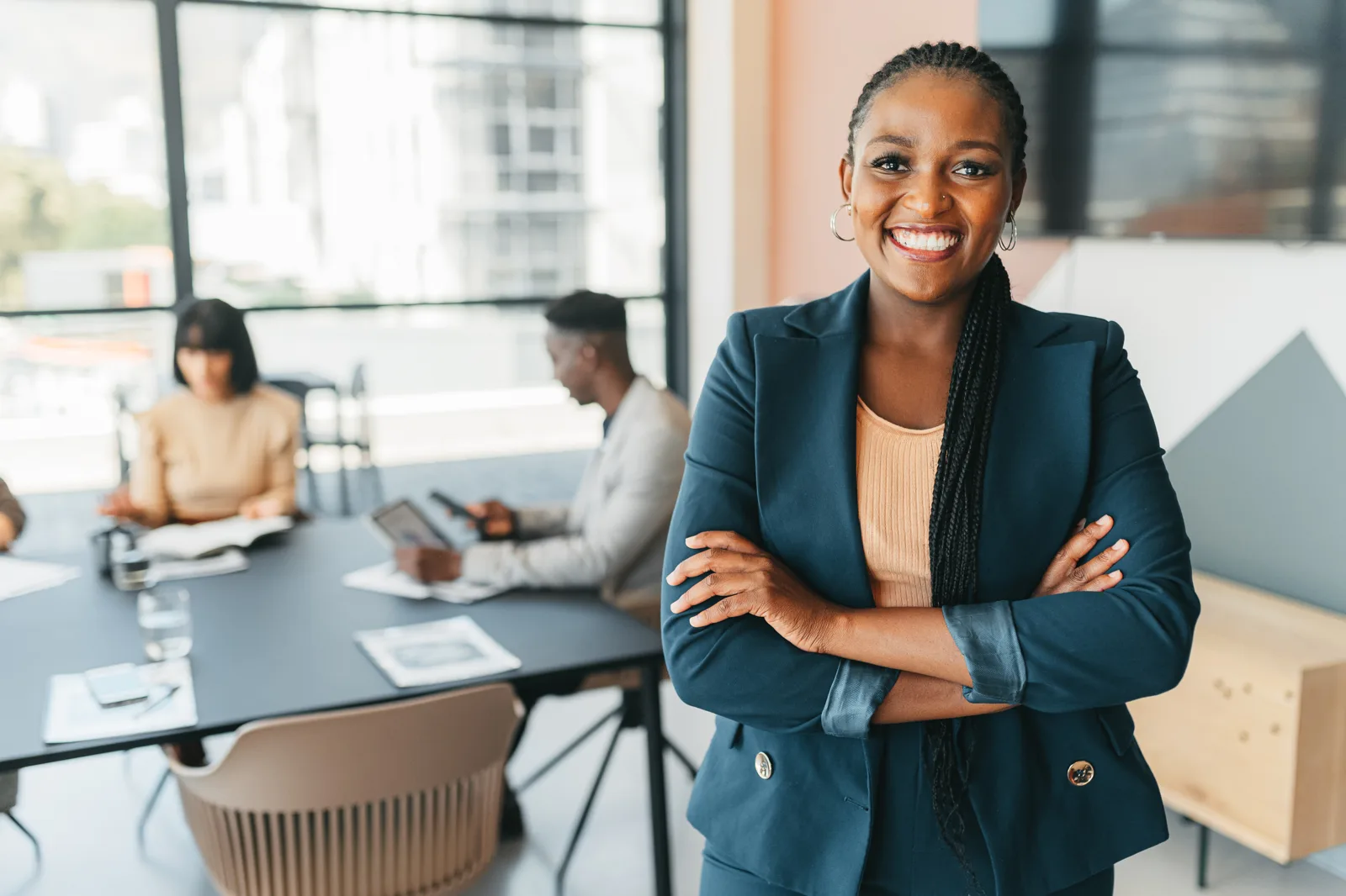 Confident business owner in a navy blazer smiling with arms crossed while her team meets in a bright office