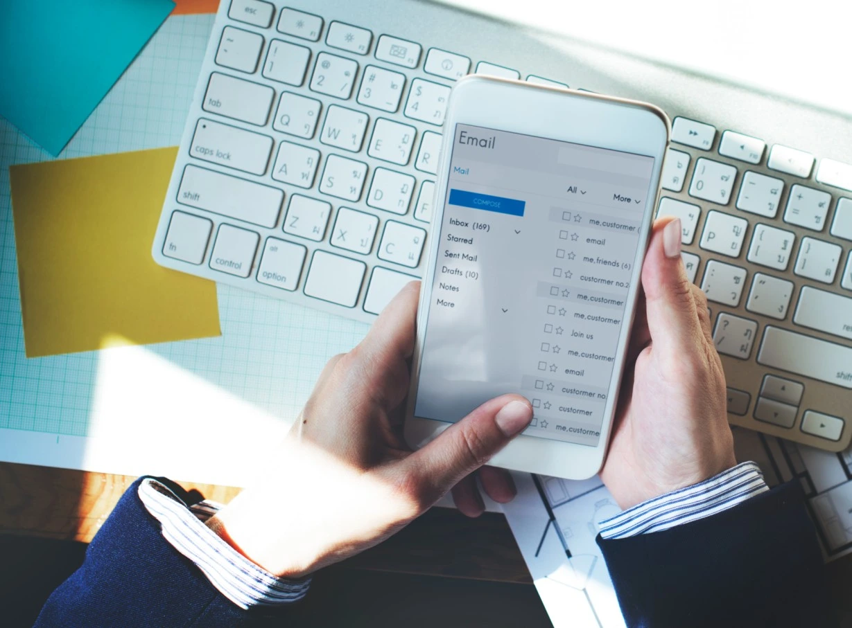 Checking Outlook email on iPhone with Mac keyboard in background, illustrating email app workflow across Apple devices