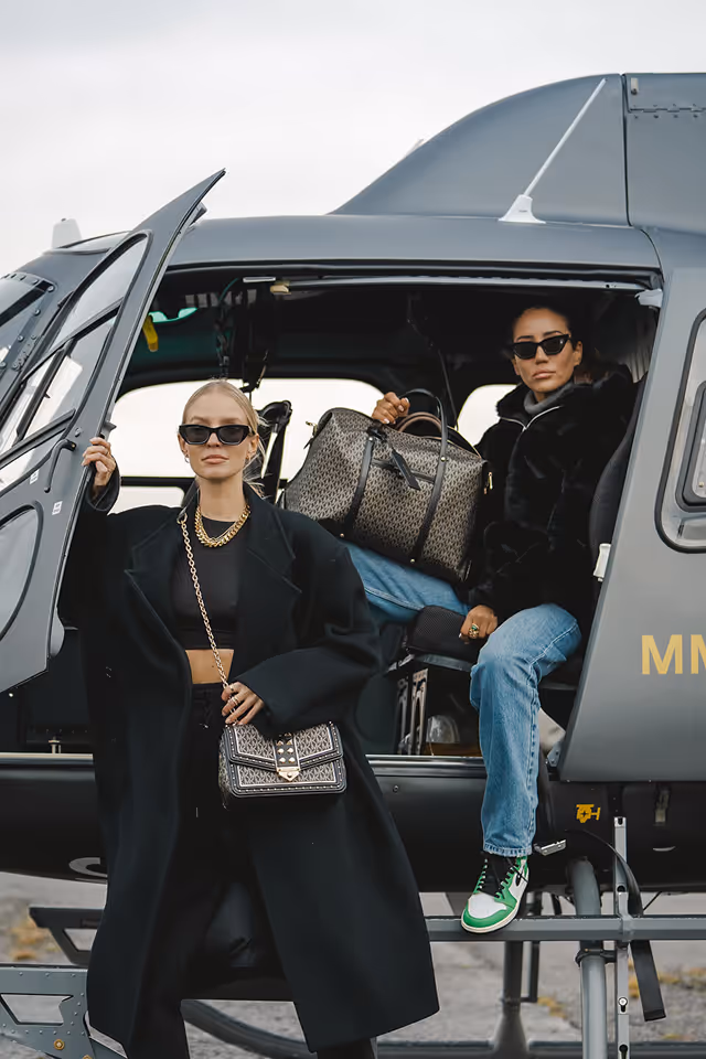 Two women wearing sunglasses and stylish coats posing with designer handbags near and inside a gray helicopter.