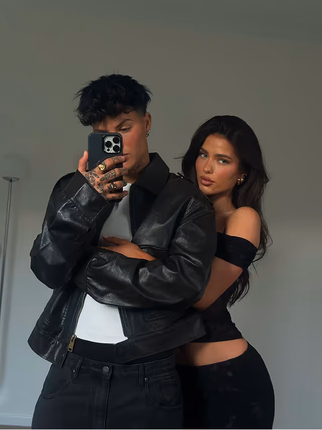 A young man in a black leather jacket taking a selfie with a smartphone while a young woman with long dark hair hugs him from behind.