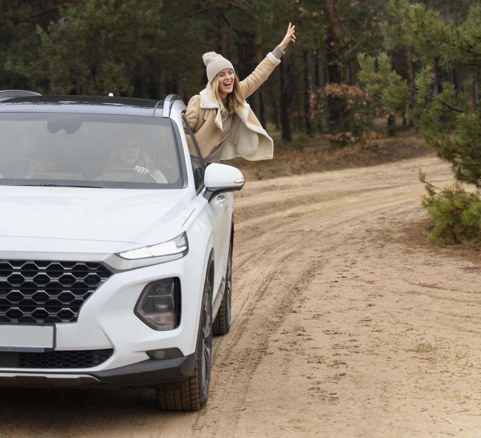 Mujer feliz con gorro saliendo de SUV blanca en camino de tierra rodeado de bosque