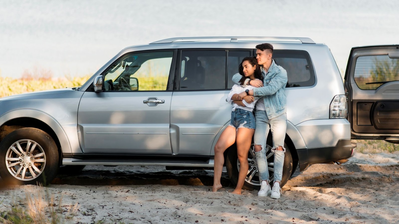 Pareja joven apoyada en SUV plateada durante viaje familiar por la playa al atardecer