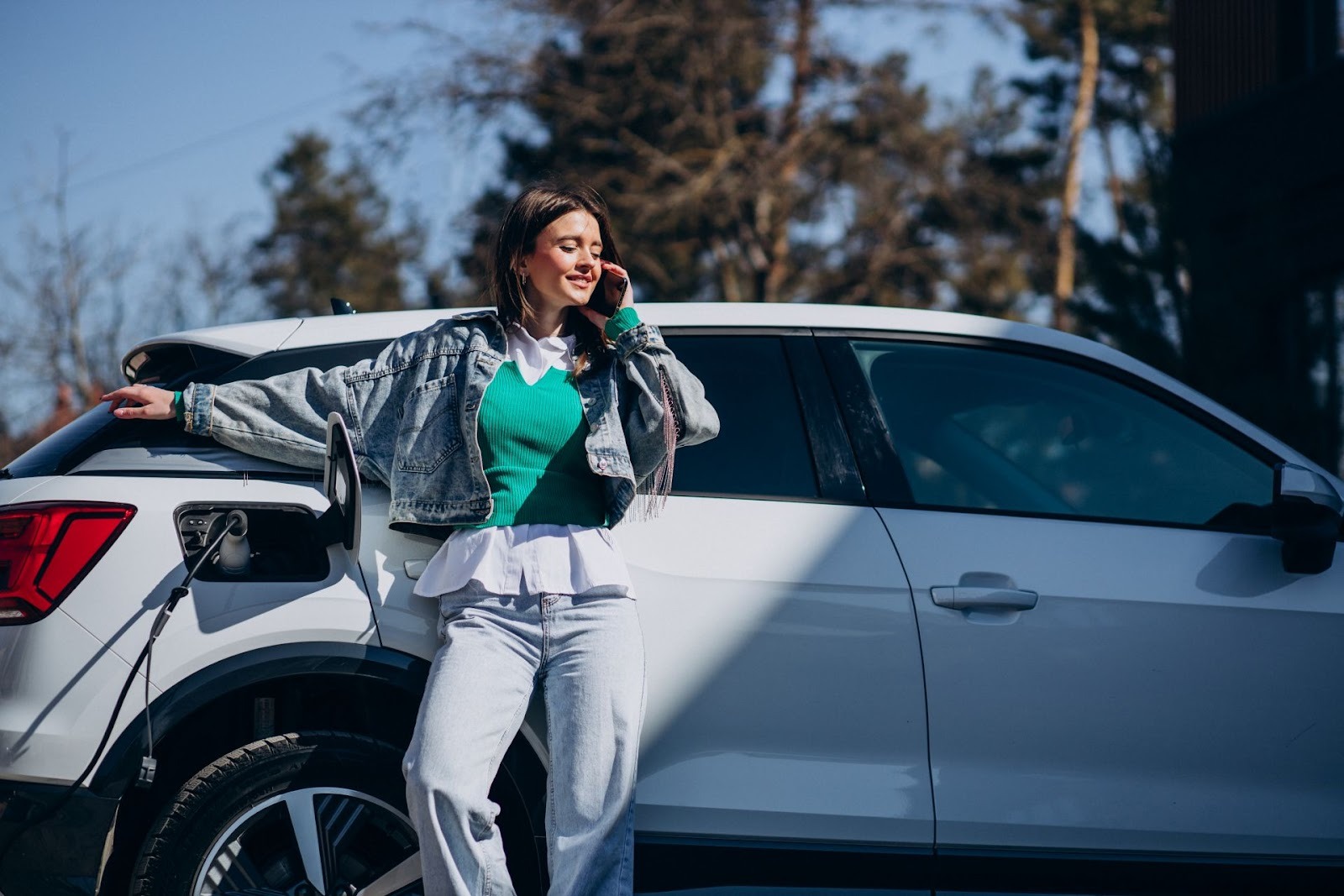 Mujer joven sonriente hablando por teléfono apoyada en SUV blanca en día soleado