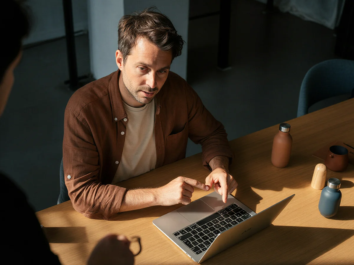 Man in brown shirt pointing at a laptop keyboard during a discussion at a wooden table.