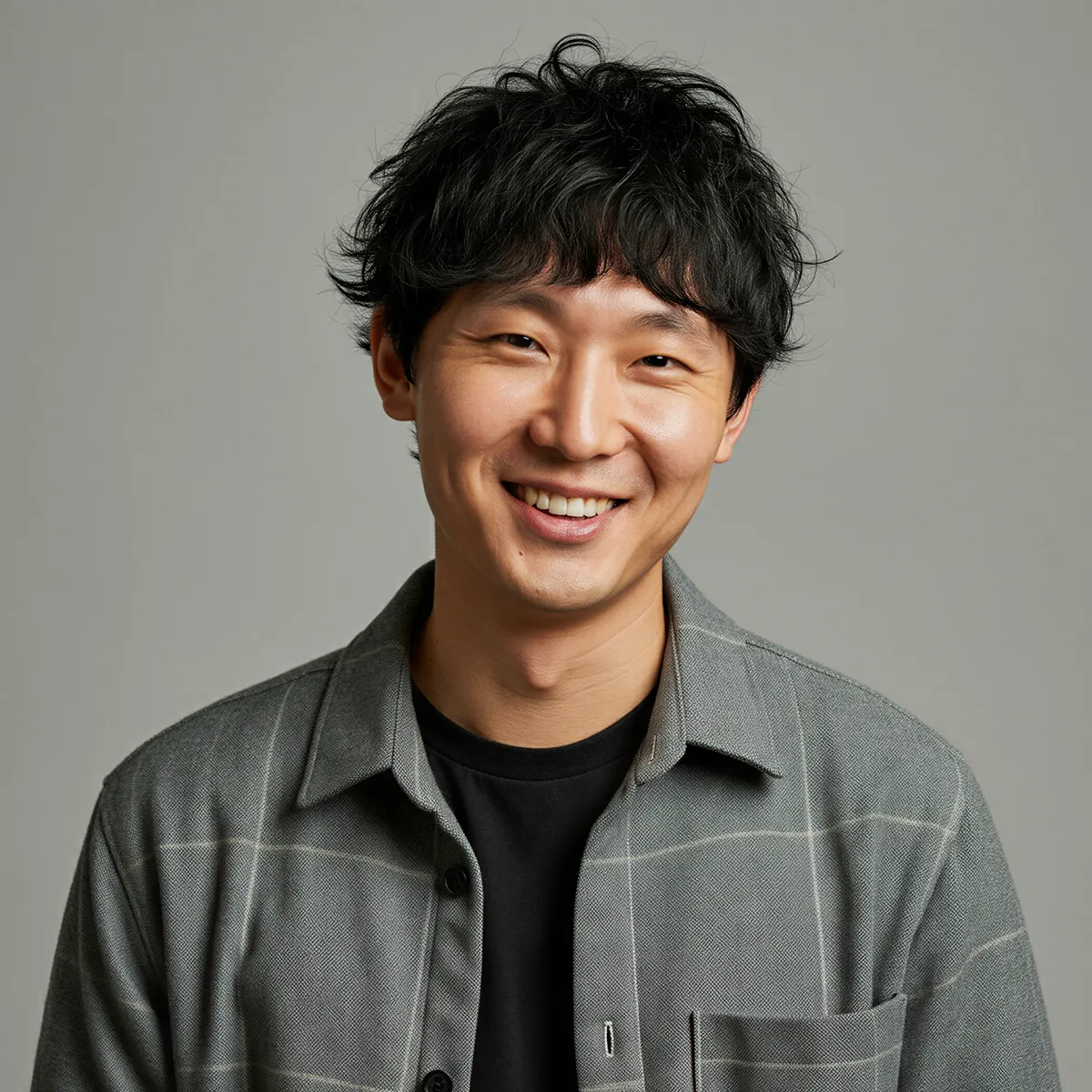 Smiling young man with curly black hair wearing a gray checkered shirt over a black t-shirt against a plain gray background.