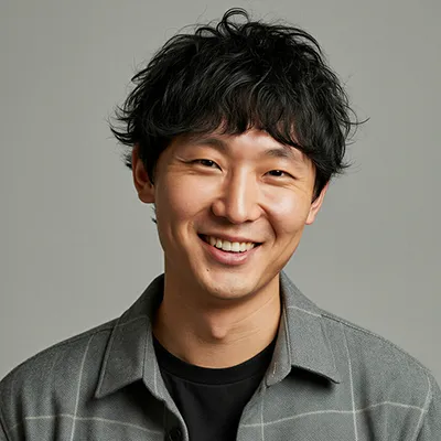 Smiling young man with black curly hair wearing a gray checkered shirt over a black t-shirt.