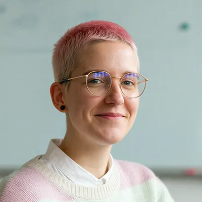 Person with short pink hair, round glasses, and a light pastel sweater smiling against a blurred background.