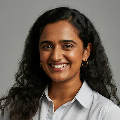 Smiling woman with long curly dark hair wearing a white shirt and gold hoop earrings against a gray background.