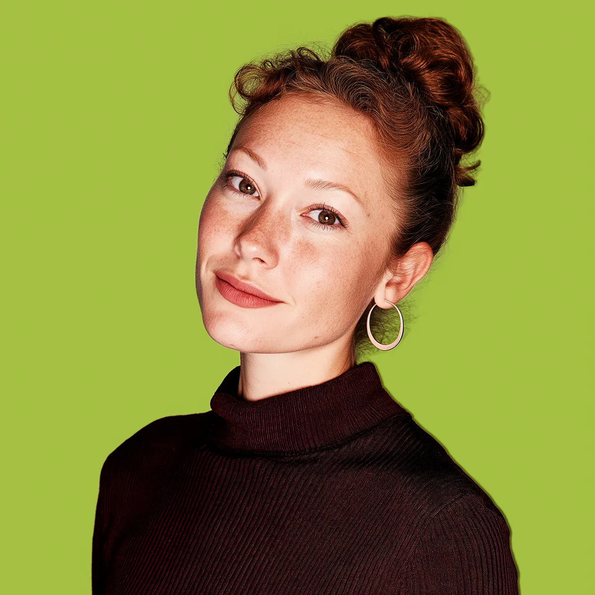 Smiling woman with curly hair in a bun, wearing hoop earrings and a dark turtleneck against a green background.