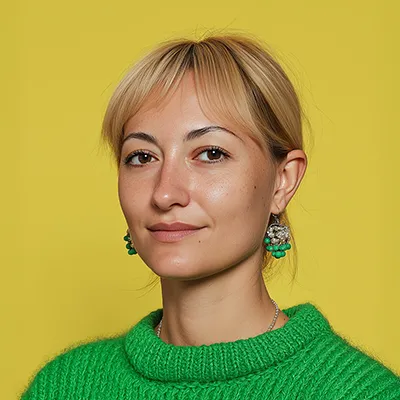 Woman with short blonde hair wearing a bright green sweater and dangling earrings against a yellow background.