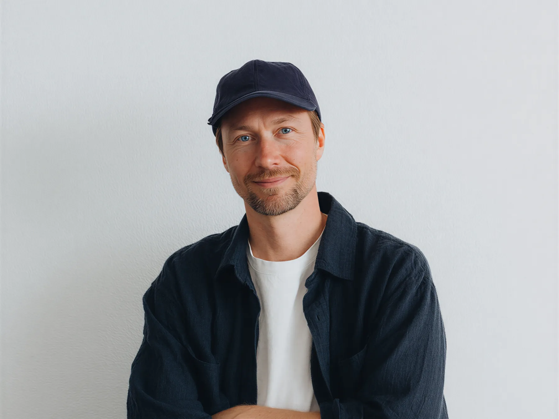 Smiling man with blue eyes wearing a black cap, black shirt, and white t-shirt against a plain light background.