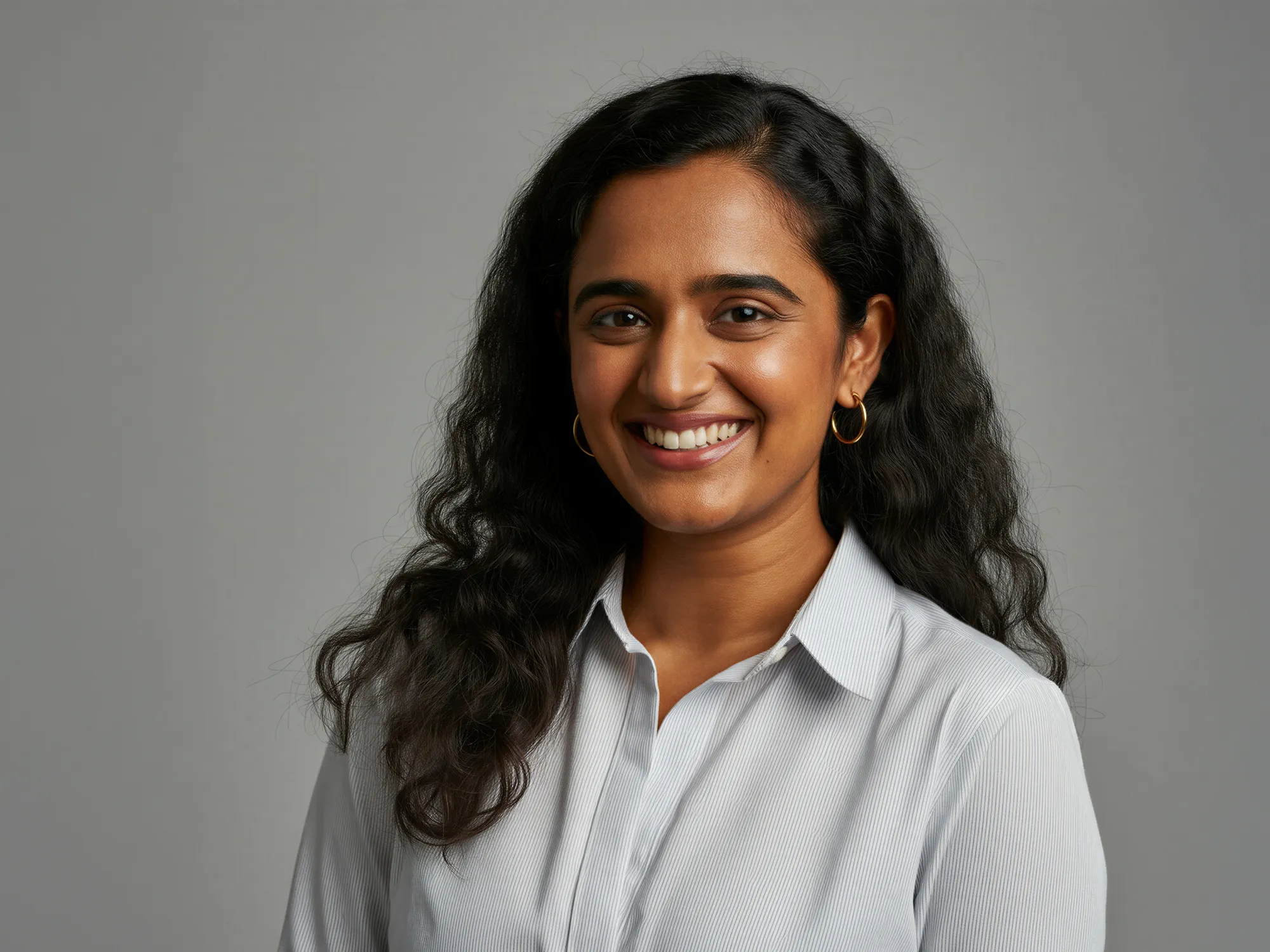 Smiling woman with long curly black hair wearing a white shirt and gold hoop earrings against a gray background.